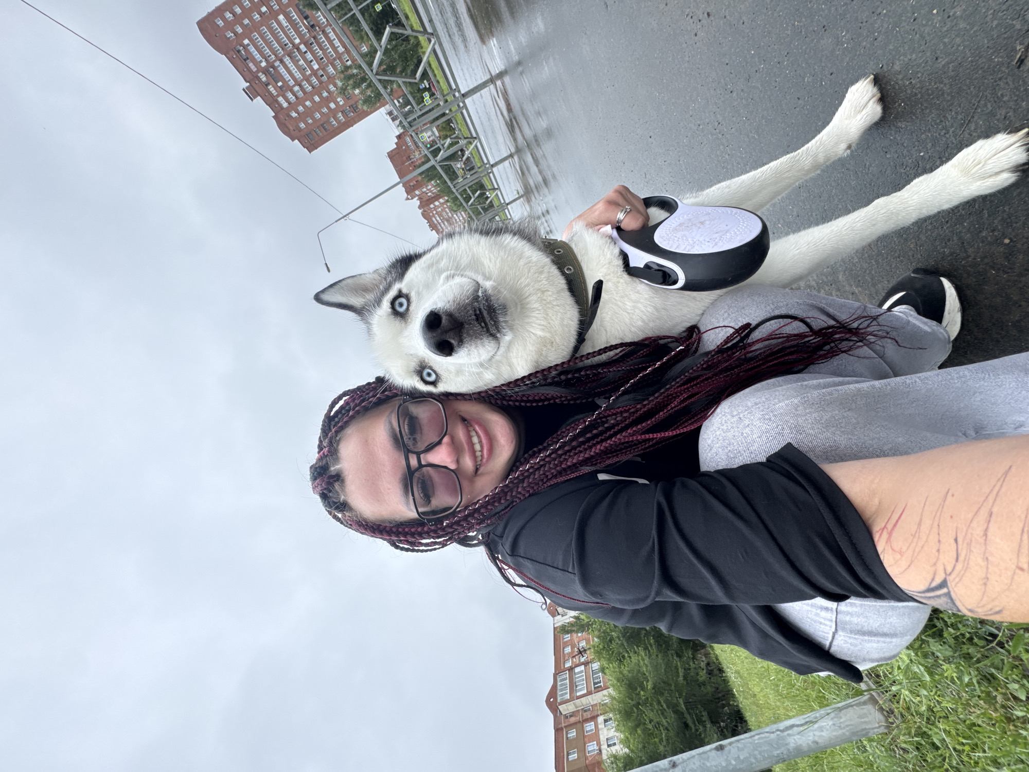 An outdoor, wide-angle, selfie-style photograph shows a young woman with long, dark red braided hair and thick-rimmed glasses, smiling and looking at the camera. She is wearing a black hooded sweatshirt and gray sweatpants. A white and black husky dog with striking blue eyes is positioned to her right, with its head tilted and looking towards the camera. The woman is holding the dog's leash, which has a black and white retractable handle. They are in an urban environment; the ground is wet, suggesting it has rained recently. In the background, there are apartment buildings, trees, a metal fence, and a streetlamp. The buildings appear to be typical of Soviet-era architecture. The presence of these buildings and the general streetscape are consistent with locations in Tolyatti, Russia.