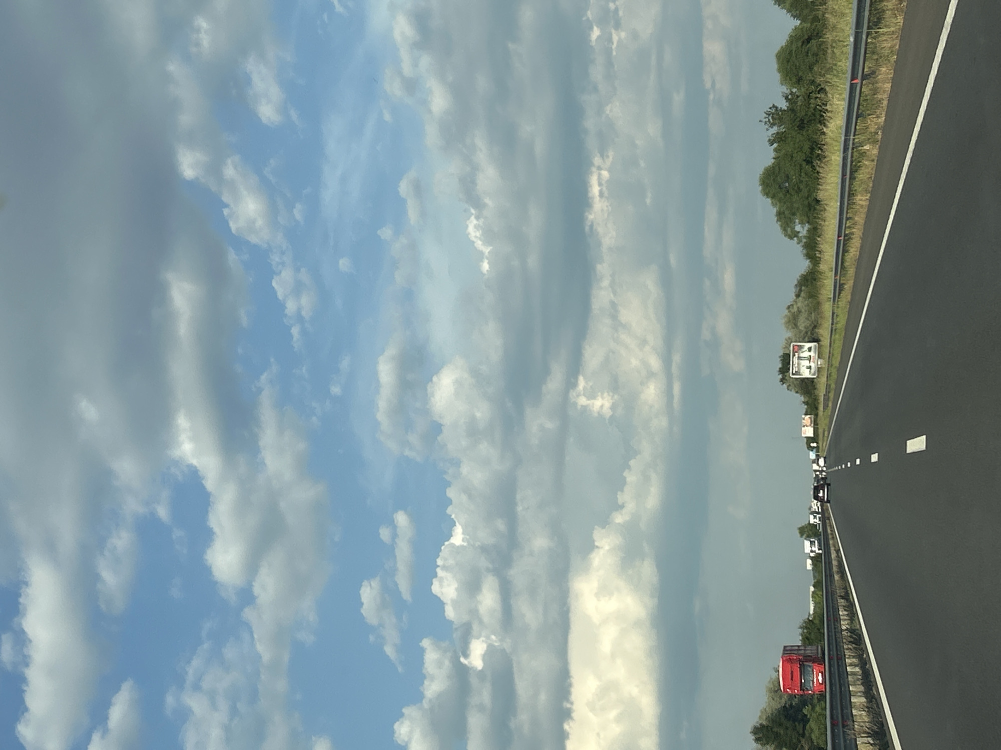 The image shows a view from a vehicle traveling on a highway. The highway is paved and has white lane markings. On the right side of the highway, there is a guardrail, grass, and trees. In the distance, there are several trucks and cars on the road. The sky is filled with clouds, and the time of day appears to be daytime. There are some billboards visible along the side of the highway, but no text is legible. The scene suggests travel or commuting, with the highway implying movement through a landscape. The presence of greenery suggests a rural or suburban setting.