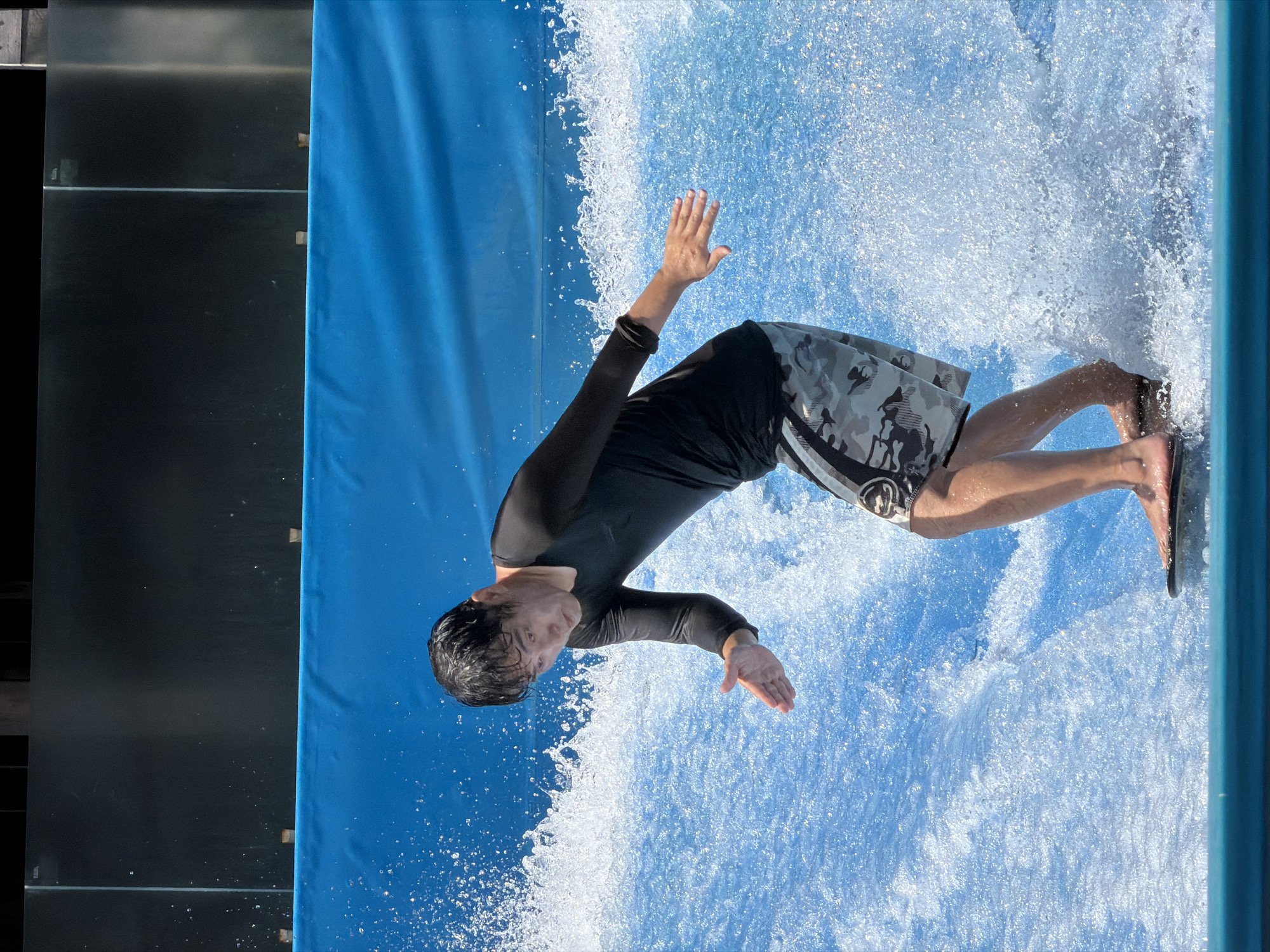A person is bodyboarding on a wave simulator with a blue backdrop and a black panel above. The person, wearing a black rashguard and camouflage shorts, is positioned on a black bodyboard, with their feet extended forward and their arms outstretched for balance. The simulated wave is creating a significant amount of splash and spray around the person. The overall scene suggests an activity related to water sports or entertainment. The location is not visible in the image.
