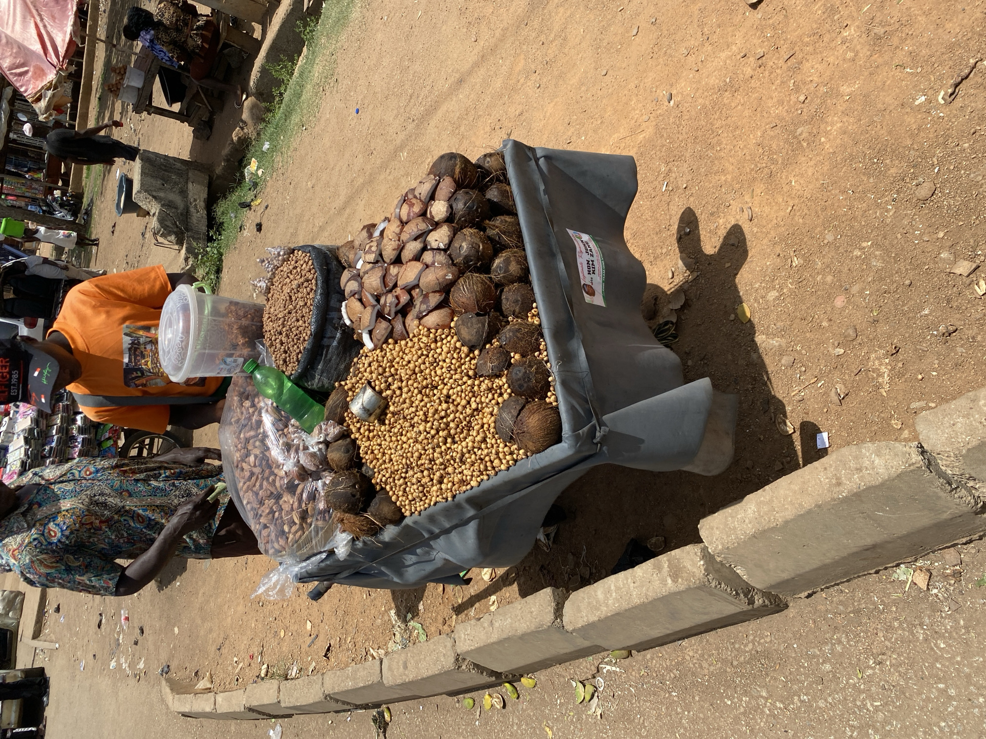 The image captures an outdoor market scene in Bukuru, Nigeria, on a bright, sunny day. The primary subjects are a vendor, partially visible in an orange t-shirt and a black cap, and a large display of goods laid out on a makeshift cart or container. Another individual in a patterned long-sleeved shirt is seen near the display, possibly a customer or assistant.
The central display holds a large quantity of goods for sale. Numerous whole coconuts and many broken coconut shells, revealing their white flesh, are piled high. Interspersed among and beneath the coconuts is a significant mound of small, round, yellowish-brown items, which appear to be tigernuts. A black sack filled with more of these round nuts is visible to the left. Additionally, a clear plastic container holds further dried goods, and a large transparent plastic bag contains elongated dried items, possibly kola nuts or similar produce. A green plastic bottle is placed near these items.
The scene takes place on unpaved, dusty ground, characteristic of an outdoor market. In the background, blurry figures and simple market stalls with wooden frames and tarpaulin covers suggest a bustling market environment. Concrete curbs delineate the edge of the selling area.
The strong, direct sunlight and short, well-defined shadows, including a distinct shadow resembling rabbit ears on the ground, indicate that it is likely midday or early afternoon on a clear day. The vendor is engaged in selling these goods.
Visible text includes "HILFIGER EST. 1985" on the vendor's black baseball cap. A white sticker on the gray tarp of the main display shows "DAN MAN NOMA" at the top, with "Aide..." and other smaller, largely illegible text beneath it, possibly a slogan or contact information.