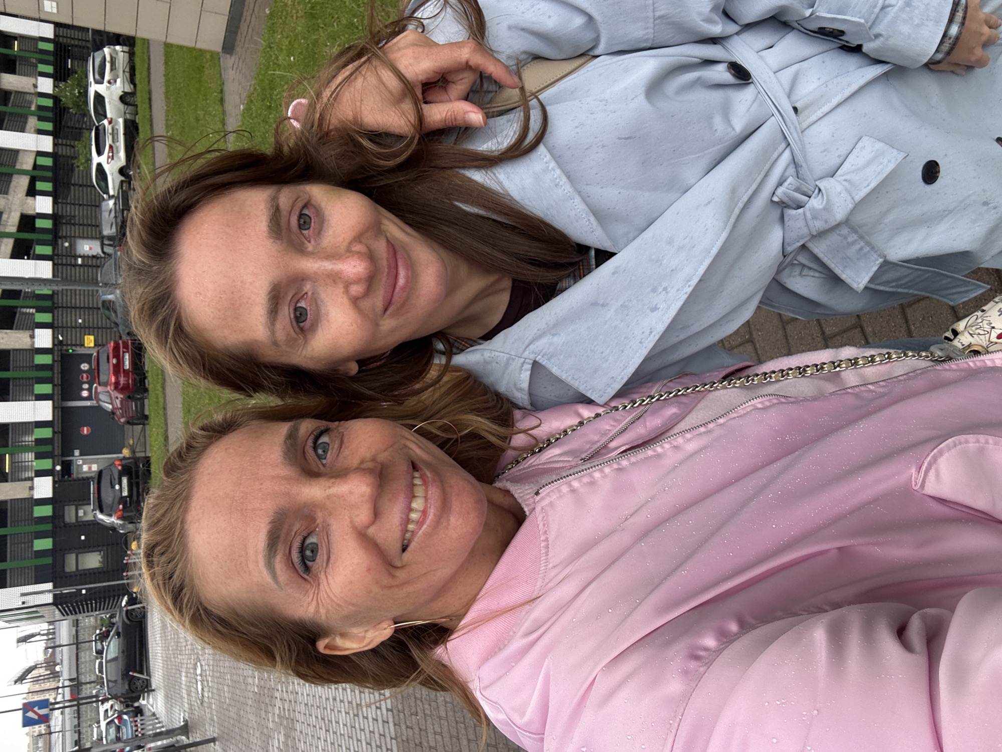 Two women are photographed close-up, with the camera pointed upwards. The woman at the bottom wears a pink jacket and hoop earrings, smiling with visible teeth. The woman above wears a light blue trench coat. Both women have freckles. They appear to be outdoors on a cloudy day. In the background, there is a multi-story building with green and black accents, and parked cars are visible. The ground is paved with light gray bricks. Based on the architectural style, the location is plausibly in Saint Petersburg, Russia.