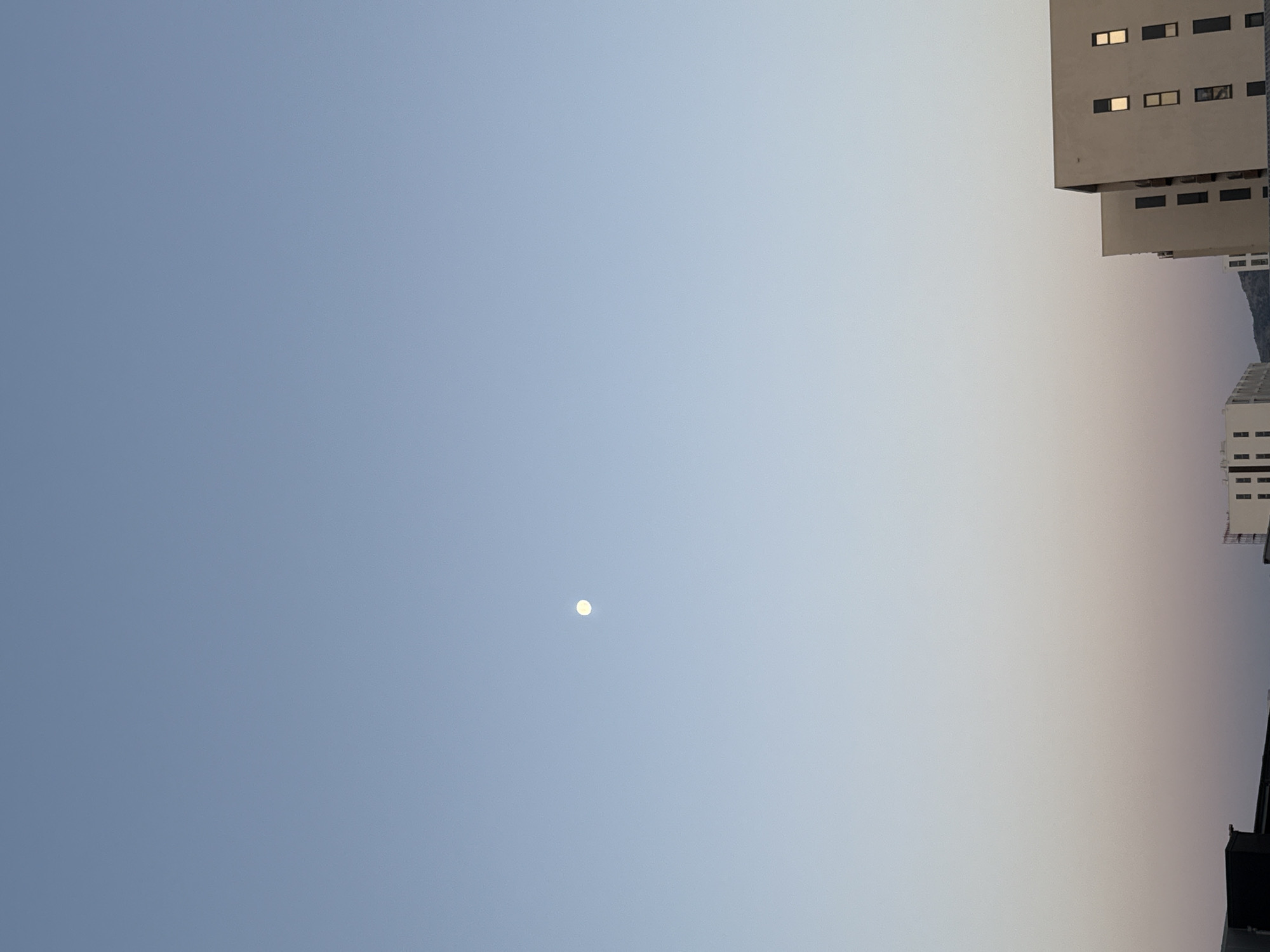 The image depicts a clear sky with a faint, pale moon visible in the upper left quadrant. The sky transitions from a light blue at the top to a soft gradient of pink and orange near the horizon. In the foreground, the silhouettes of buildings are visible against the sky. The buildings appear to be modern apartment or office blocks, with some windows reflecting the light. The overall scene suggests an urban environment at either dawn or dusk. There are no people or animals visible in the image. The location is an unknown city.
