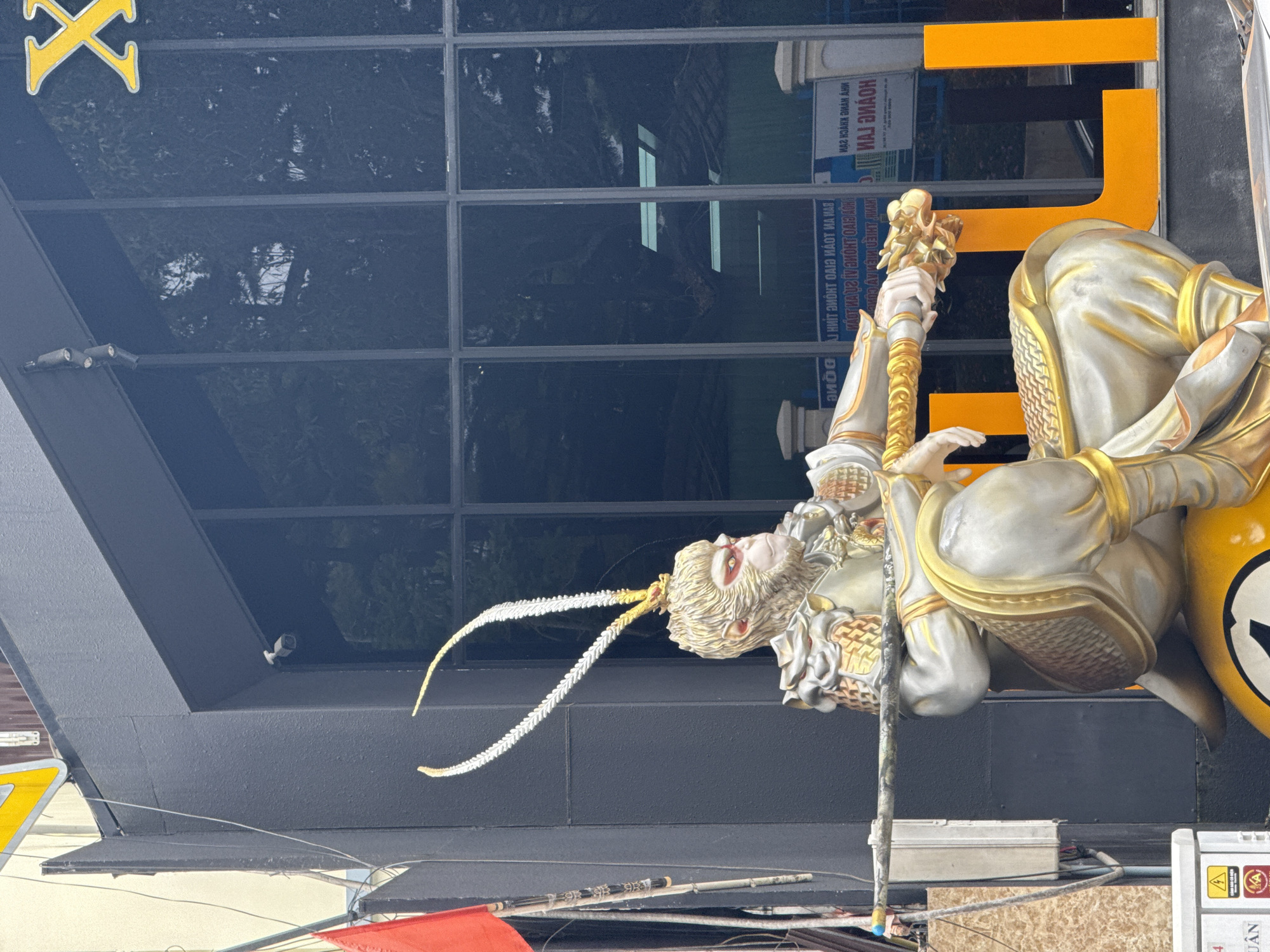 A statue of a character resembling the Monkey King from Journey to the West, with intricate golden armor and distinctive headdress, is positioned prominently in front of a building with a dark, reflective facade. The statue is seated, holding a staff horizontally across its lap. Behind the statue, the building's large, dark windows reflect the surrounding environment, including trees and some interior lighting. Large, orange, stylized letters, partially obscured by the statue, are visible on the right side of the image. A red flag is mounted on a pole to the left of the statue. The scene appears to be on a street in Da Lat, Vietnam, possibly in front of a business or establishment. Electrical wires and a security camera are also visible on the building's exterior.