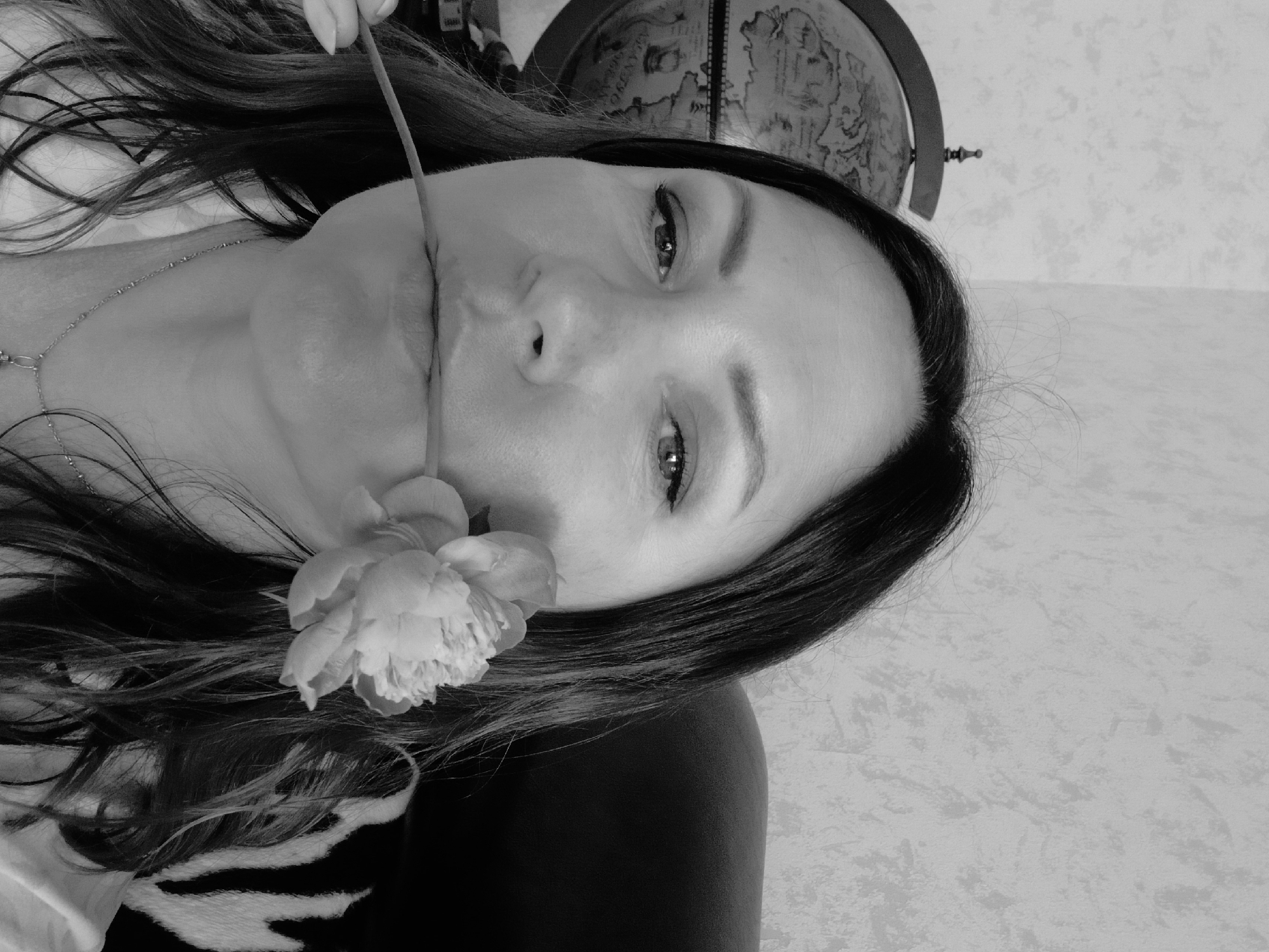 This is a black and white, low-angle, close-up photograph of a woman's face. She is lying down, and the camera is positioned above her. A thin stem, holding a light-colored flower, is placed between her lips, obscuring her mouth. Her eyes are open and looking directly at the camera, with a slightly melancholic or thoughtful expression. Her long, dark hair is spread out around her head. A delicate silver necklace is visible around her neck. In the background, a portion of a globe with a detailed map is visible, positioned behind and to the left of her head. The wall behind the globe has a textured, light-colored pattern. The lighting appears to be soft and diffused. There is no discernible time of day, weather, or specific geographical location information.