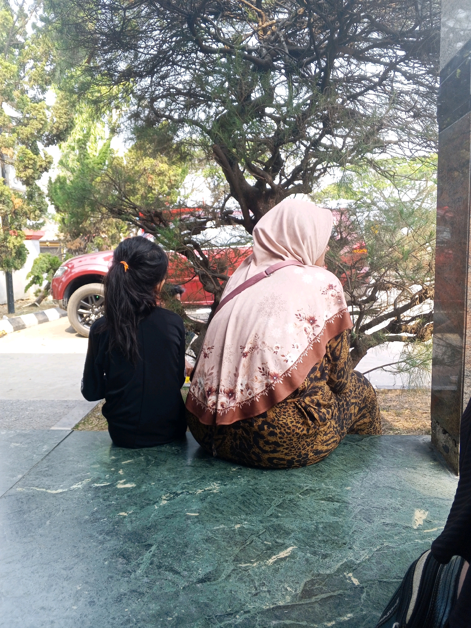 The image depicts two individuals seated on a green, marble-like surface. The person on the left has long, dark hair tied in a ponytail and is wearing a black long-sleeved shirt. The person on the right is wearing a light-colored hijab with a floral pattern and a patterned dress with a leopard print. They are seated outdoors, with a large, gnarled tree behind them. In the background, a red vehicle is visible, along with some buildings and foliage. The overall setting appears to be a public space, possibly a park or a courtyard, in Ciruas, Indonesia. The lighting suggests it is daytime, with sunlight filtering through the trees.