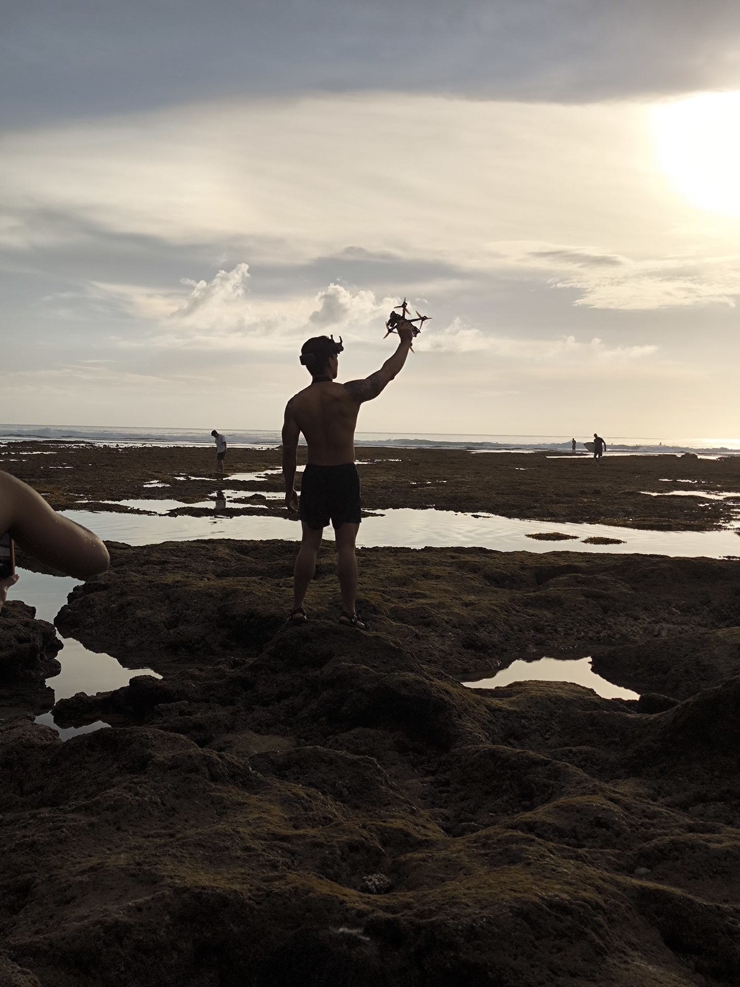 A partially silhouetted man stands barefoot on a rocky, uneven terrain at low tide, raising a drone overhead. The man wears shorts, a head-mounted device, and appears to have a tattoo on his left arm. The rocky landscape is interspersed with shallow pools of water reflecting the sky. In the distance, the ocean meets the horizon under a partly cloudy sky with the sun breaking through. Other individuals are visible further out on the terrain. The location is Uluwatu, Indonesia.