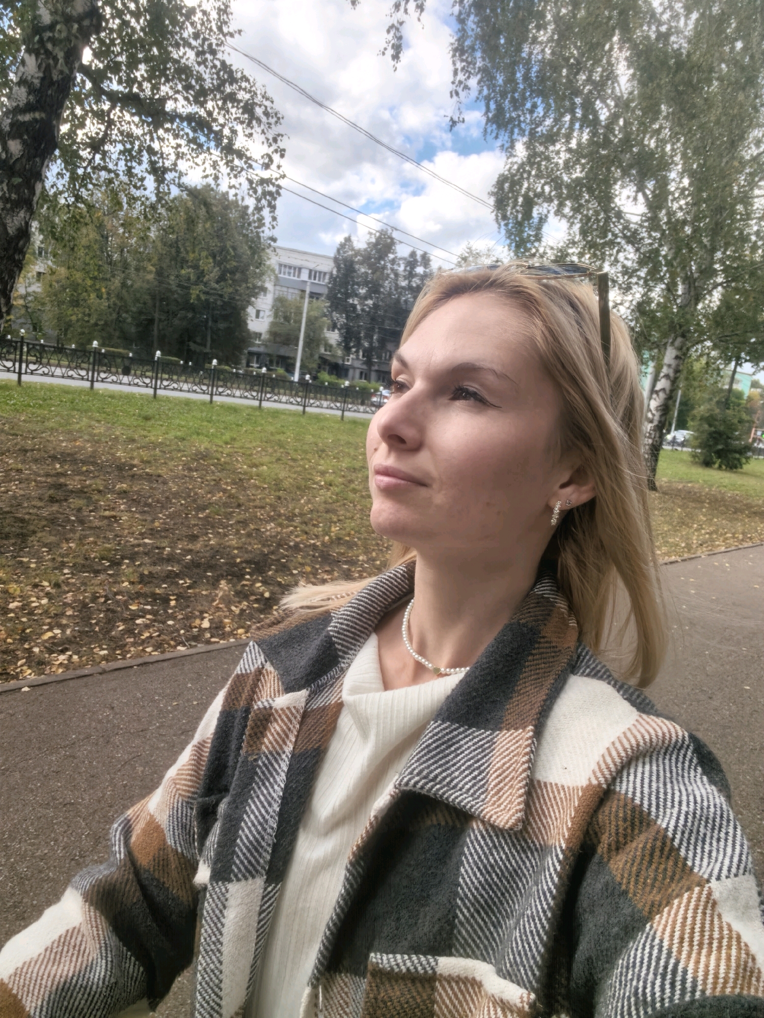 A mid-shot, taken from a low angle, features a blonde woman standing on a paved path next to a grassy area with fallen leaves. She is wearing a cream-colored sweater and an oversized plaid jacket in shades of grey, white, and brown. A pearl necklace with a gold pendant is visible around her neck. She has blonde hair and is looking up and to her right with a contemplative expression. Sunglasses are perched on top of her head.
In the background, a black wrought-iron fence separates the path from a grassy embankment. Beyond the fence, a row of trees with green and yellowing foliage lines a street. In the distance, a multi-story building with many windows is visible. The sky is partly cloudy with patches of blue. The scene appears to be in a park or a residential area in the city of Sterlitamak, Russia, judging by the architecture and vegetation.