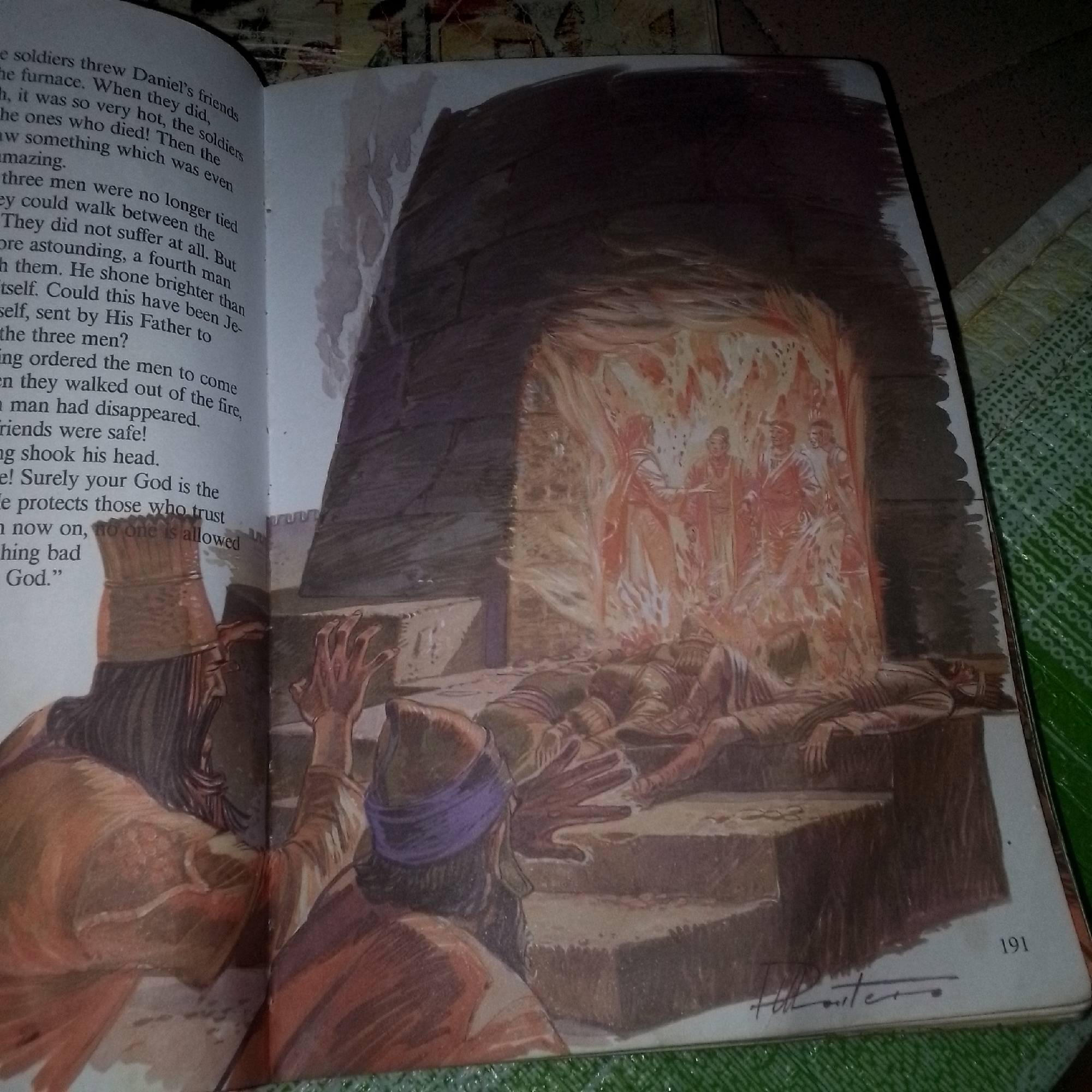 The image depicts an illustration from a book with text on the left page and a painting on the right. The text describes friends of Daniel being thrown into a furnace, which was very hot, and that they did not suffer. It mentions a fourth man, sent by their Father, who shone brighter than themselves. The painting illustrates this scene, showing three men lying on the ground, seemingly unharmed, in front of a fiery furnace. Inside the furnace, three figures are visible, surrounded by flames, with a fourth figure standing among them. Two figures are seen outside the furnace on the left, one wearing a tall headdress and robes, and another with a purple band around his head, gesturing with raised hands. The scene takes place outdoors, with stone structures in the background. The right page also shows the number "191" and a signature.