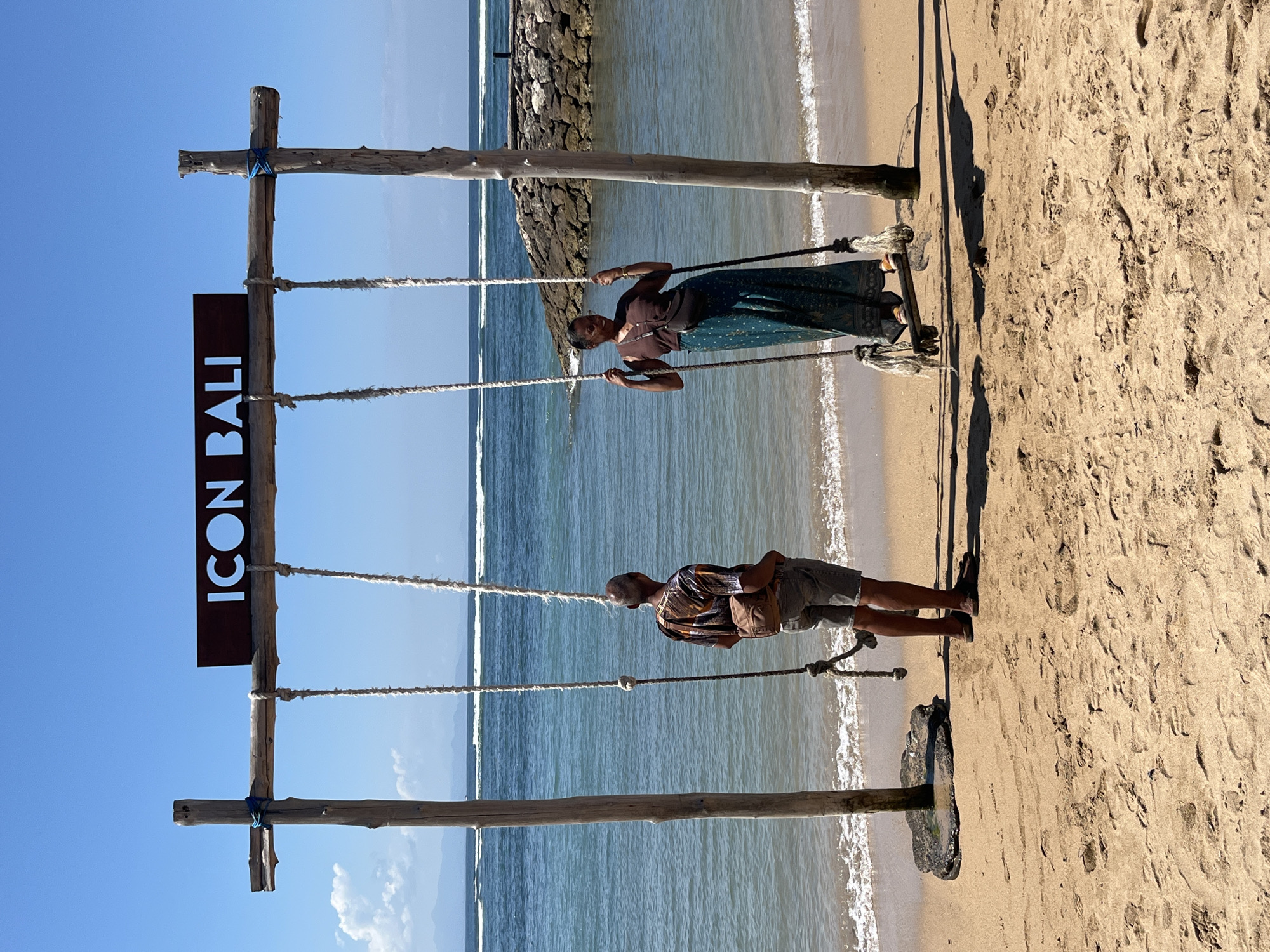 Two individuals are on a sandy beach in Denpasar, Indonesia, near a swing set. On the left, a person wearing a patterned short-sleeved shirt and shorts stands facing away from the camera, looking out at the ocean. They have a brown fanny pack slung across their chest. On the right, a person wearing a brown short-sleeved shirt and a long, patterned skirt is seated on a swing, holding onto the ropes. They are looking towards the left, with a slight smile.
The swing set consists of two wooden posts supporting a horizontal wooden beam. A dark sign with white lettering spelling "ICON BALI" is mounted on the beam. The swings are made of wooden seats suspended by thick ropes. The ocean is visible in the background, with gentle waves lapping at the shore. A rocky structure extends into the water on the right side of the frame. The sky is clear and blue. The sand is light brown and shows footprints.