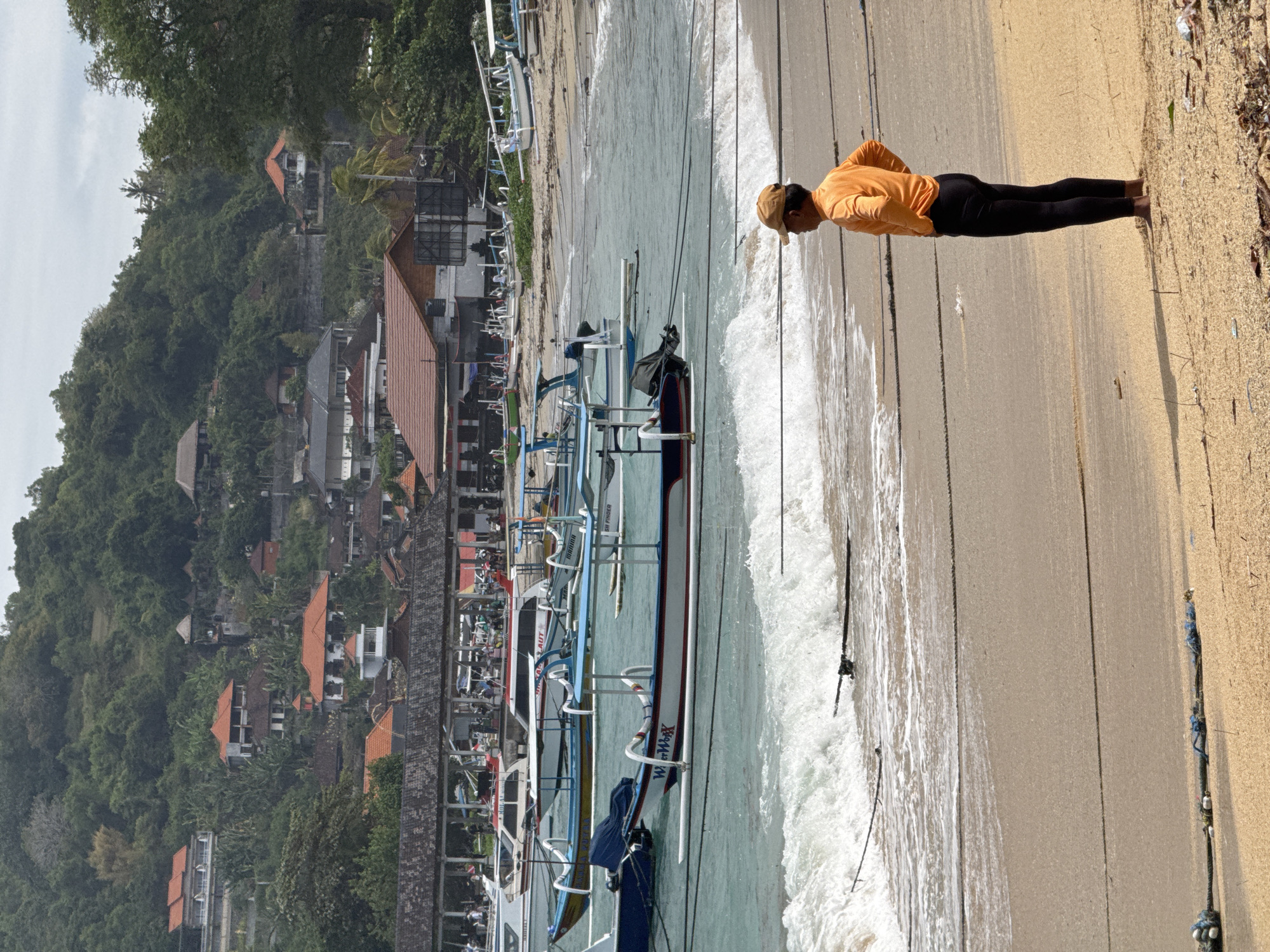 In Manggis, Indonesia, this image captures a beach scene with a man in an orange shirt and black pants standing on the sand, facing the ocean. His hands are on his hips, and he wears a tan baseball cap. The ocean is visible with moderate waves breaking on the shore. Numerous traditional Indonesian fishing boats, known as jukungs, are anchored in the shallow turquoise water, some with their outriggers visible. In the background, a densely vegetated hillside is dotted with buildings, featuring orange and brown tiled roofs, suggesting a residential or resort area. A covered walkway or pier is situated along the shoreline. The beach itself is sandy, with scattered debris near the foreground. The sky is overcast.