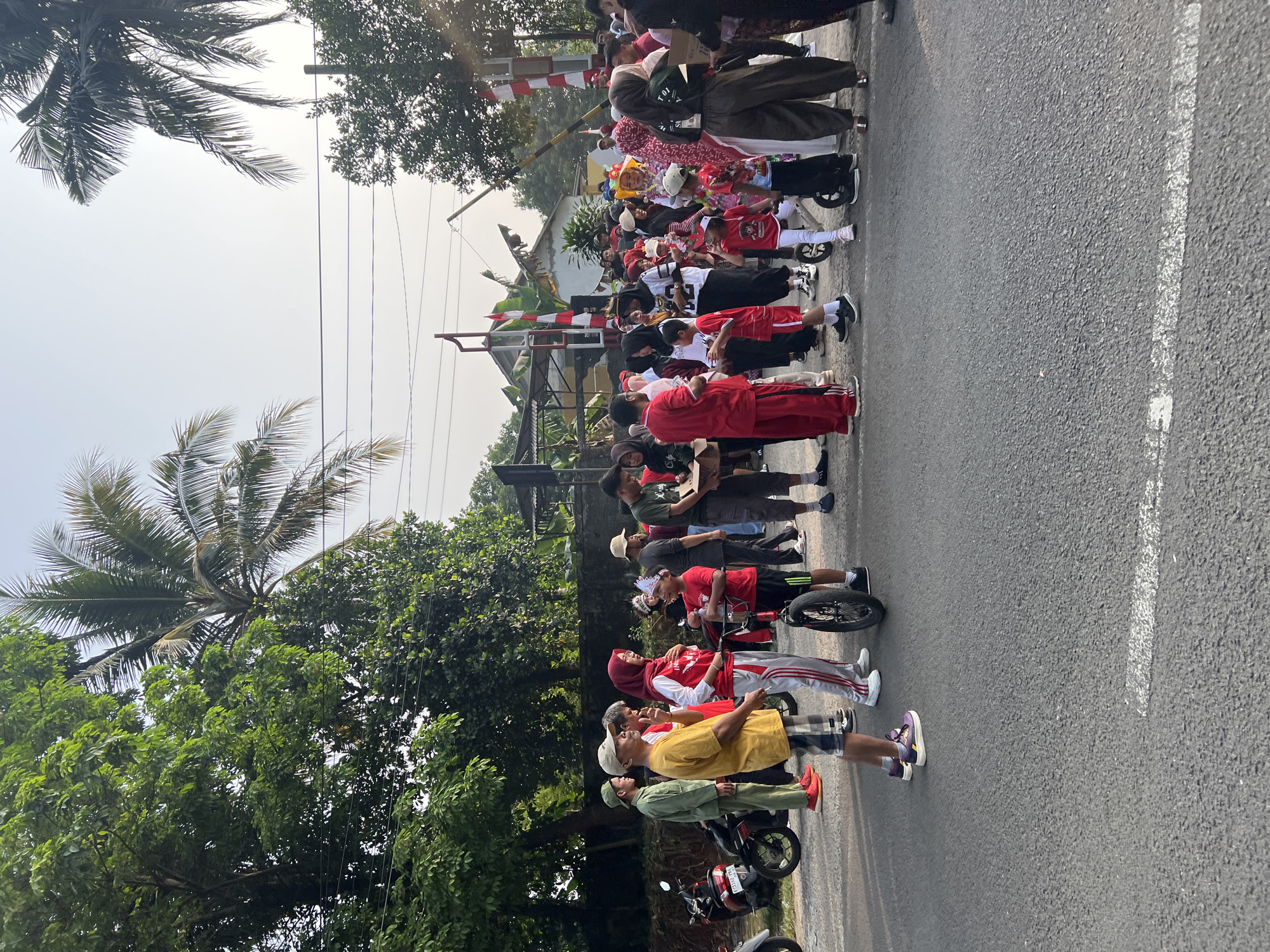 In Cibinong, Indonesia, a crowd of people, many dressed in red and white, have gathered on a paved road, potentially for a local event or celebration. Palm trees and lush green foliage line the background, with power lines stretching across the frame. Several motorcycles are parked on the side of the road. The people are of various ages, with adults and children intermingling. Some individuals are holding boxes, suggesting they may be distributing items. A small bicycle with training wheels is present, with a child standing next to it. The overall atmosphere appears to be one of community gathering.