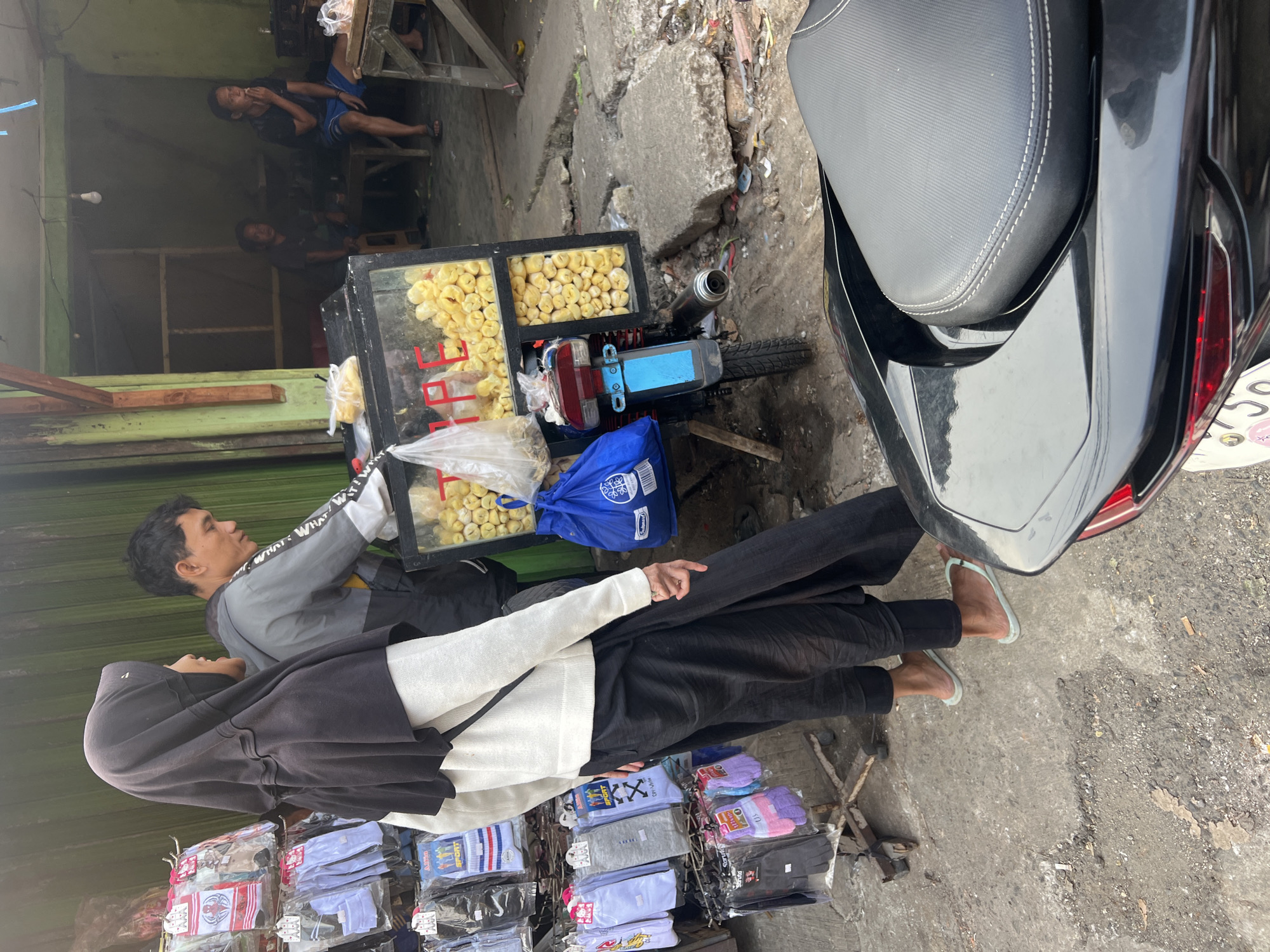 In Citeureup, Indonesia, a street vendor operates a food cart attached to a motorcycle. The cart is a glass-fronted display filled with what appear to be yellow fried snacks. A man, likely the vendor, is visible interacting with the cart. Two women stand to the left of the cart. One, wearing a black hijab and a white top, faces the vendor. The other, partially obscured, is also looking towards the vendor. Racks of socks and gloves are displayed to the left of the women. The background shows a simple building with a green facade and a wooden structure. Two men are seated inside the building in the background. The ground is paved with concrete and is uneven. A black motorcycle seat is prominently in the foreground.