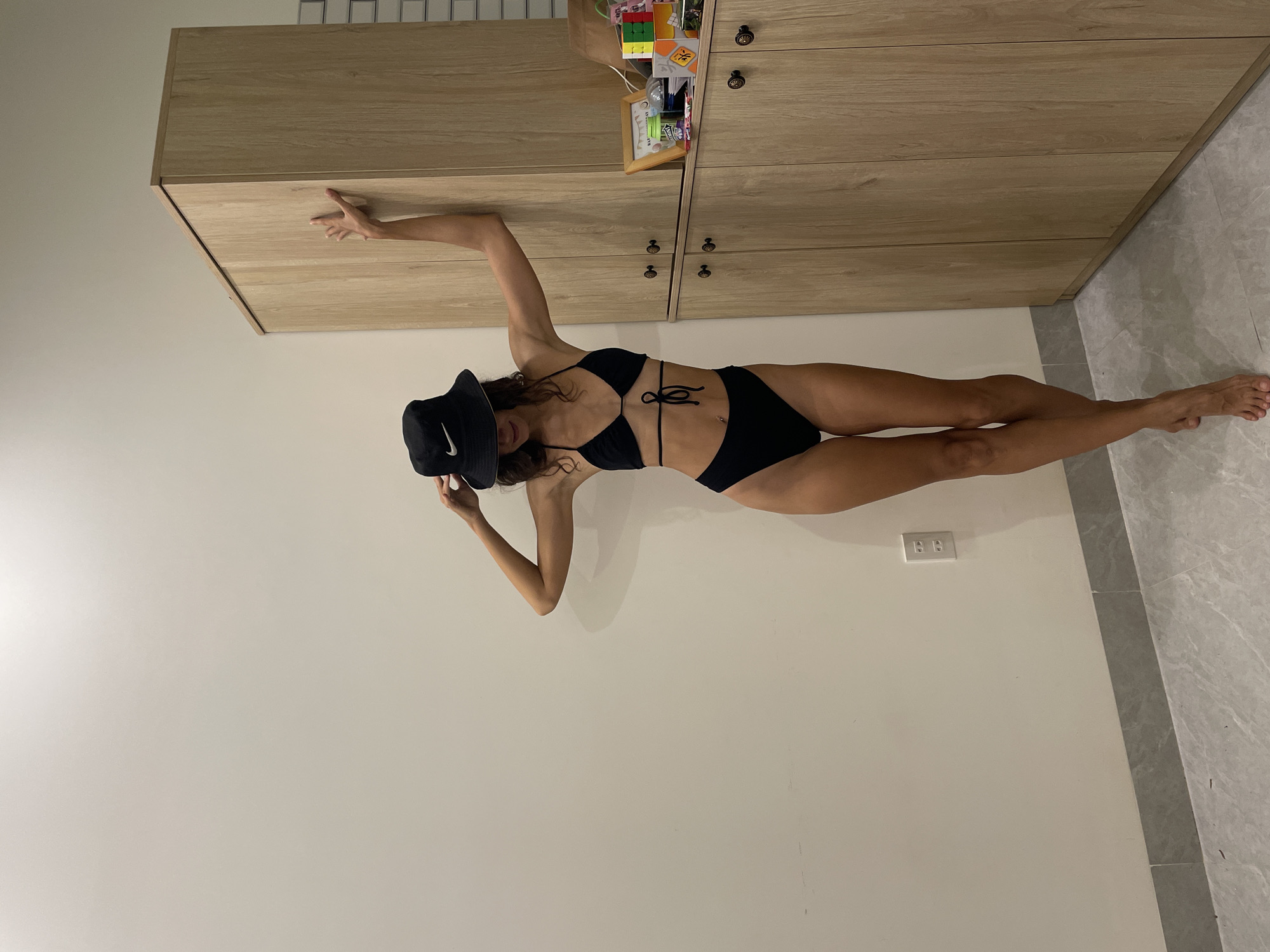 An upright, full-body shot features a woman posing indoors, wearing a black two-piece bikini and a black bucket hat with a white Nike logo. Her face is partially obscured by the hat, which she holds with her left hand. Her right arm is extended upwards, with her palm resting against a light-colored wooden cabinet.
The setting appears to be a modern indoor room, possibly in Mũi Né, Vietnam. Behind the woman are several wooden cabinets or drawers with small, dark handles. On top of the upper cabinet, various small decorative items are visible, including a Rubik's cube and a framed picture. The wall to the woman's left is plain and light-colored, while the floor consists of light gray tiles. An electrical outlet is visible on the wall. The woman is standing on the tiled floor. The overall impression is a casual, posed portrait in a well-lit indoor environment.
