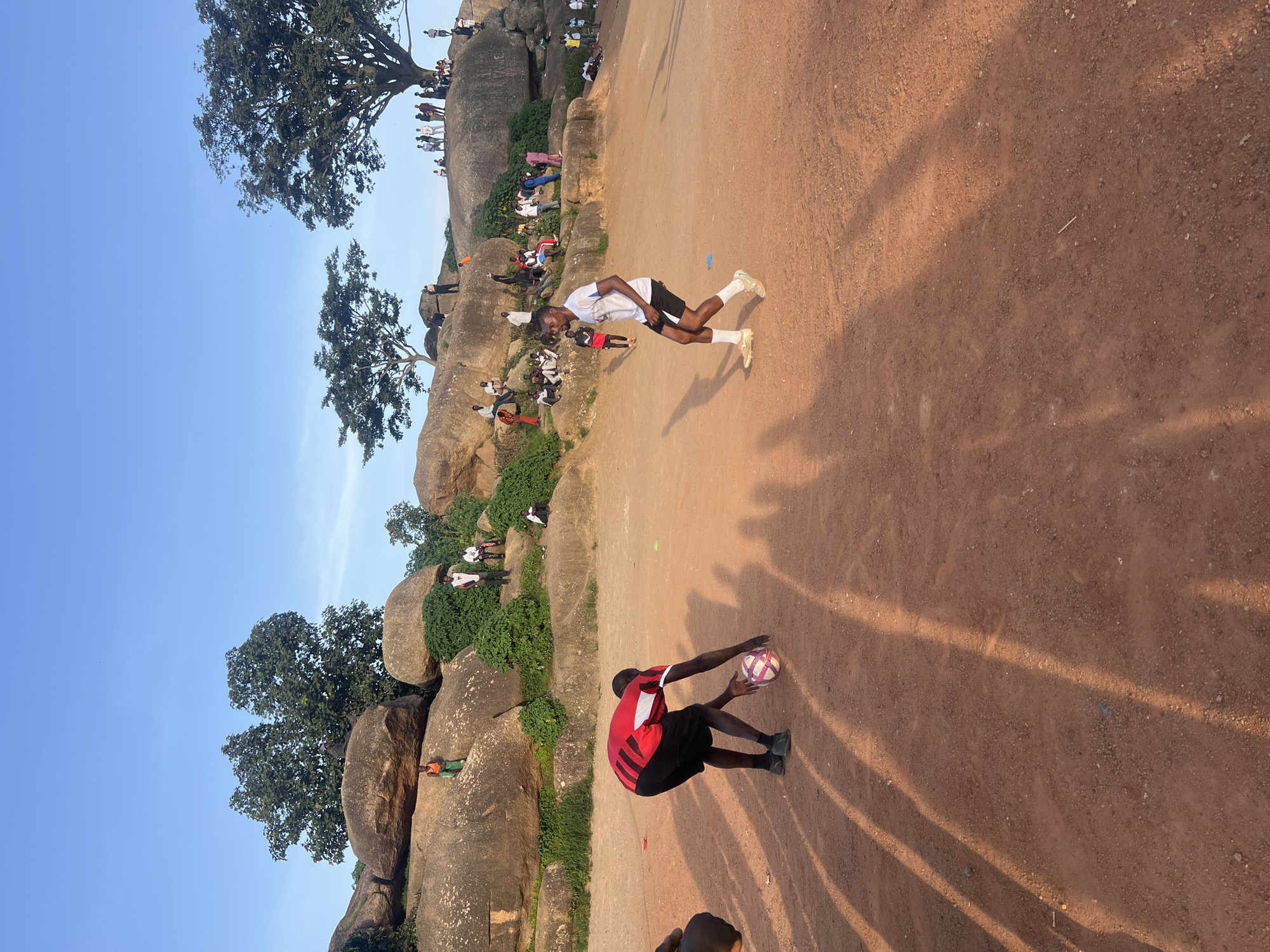 The image depicts a group of people in an outdoor, sandy area with large rock formations in the background. Two young men are actively engaged in a game of soccer in the foreground. One, wearing a red and black striped shirt and black shorts, is bent down, reaching for a soccer ball. The other, in a white t-shirt and black shorts, is standing, facing away from the viewer, seemingly in the process of playing. Numerous other people are scattered throughout the scene, some sitting on the rocks and others standing, observing the activity. The environment appears to be a natural landscape, possibly a park or recreational area, with sparse vegetation and clear blue sky overhead. The lighting suggests it is late afternoon or early evening, casting long shadows across the ground. This scene is characteristic of recreational spaces found in the city of Jos, Nigeria, known for its rocky topography and vibrant outdoor activities.