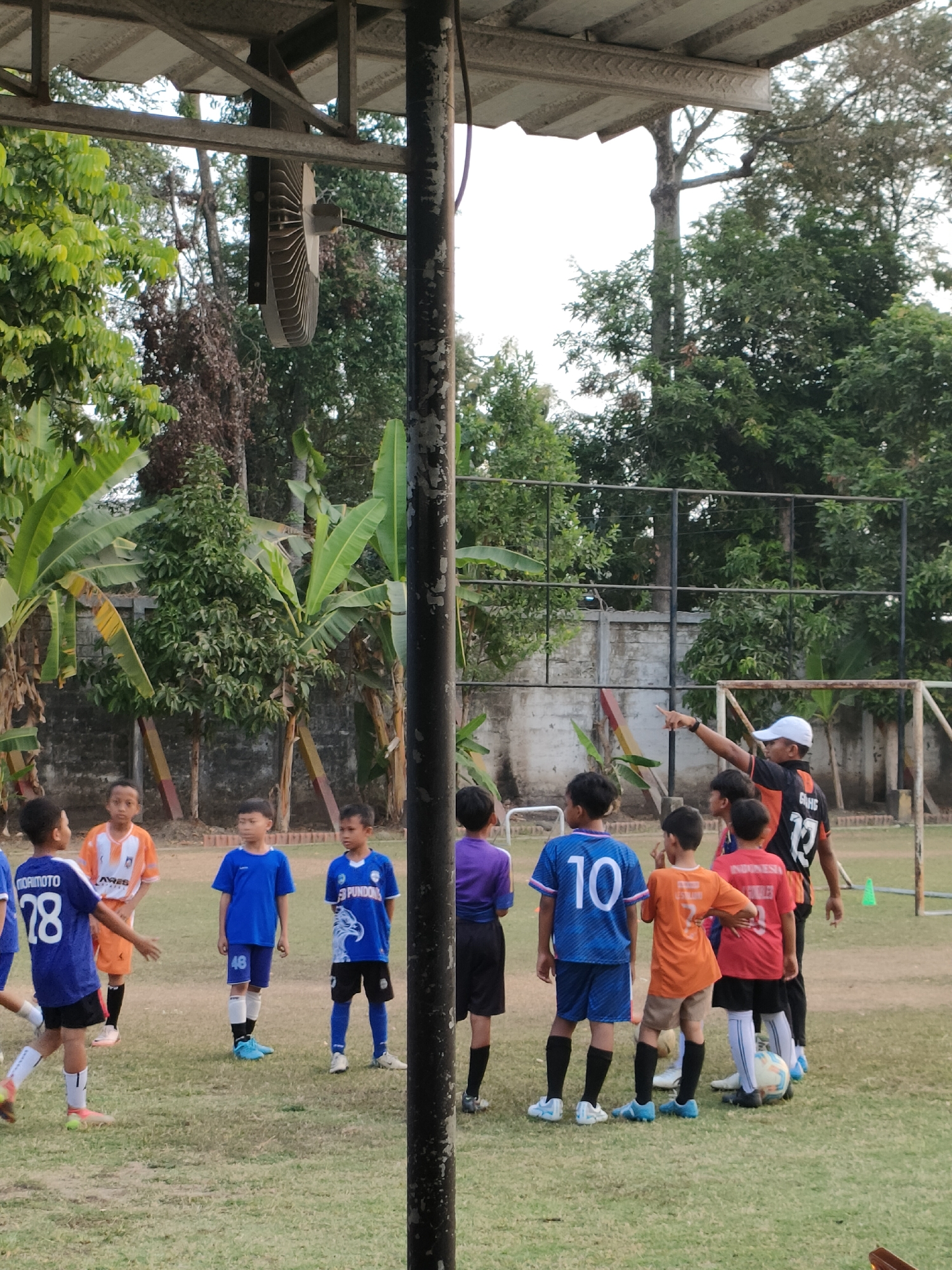 The image depicts a group of young boys in soccer uniforms on a grassy field, likely a training session or casual game. A coach, wearing an orange and black jersey and a white cap, stands facing the boys and points with his right hand. The boys are varied in age and are clustered together, some looking at the coach, others at each other. One boy in a blue jersey with "MORIMOTO 78" is in motion. A soccer ball is visible near the feet of a boy in a red jersey. The background shows lush green trees, a concrete wall, and part of a soccer goal. The presence of a large industrial fan mounted overhead suggests this might be an outdoor sports facility or park. The general setting indicates a tropical or subtropical climate. The location is specified as Banguntapan, Indonesia.