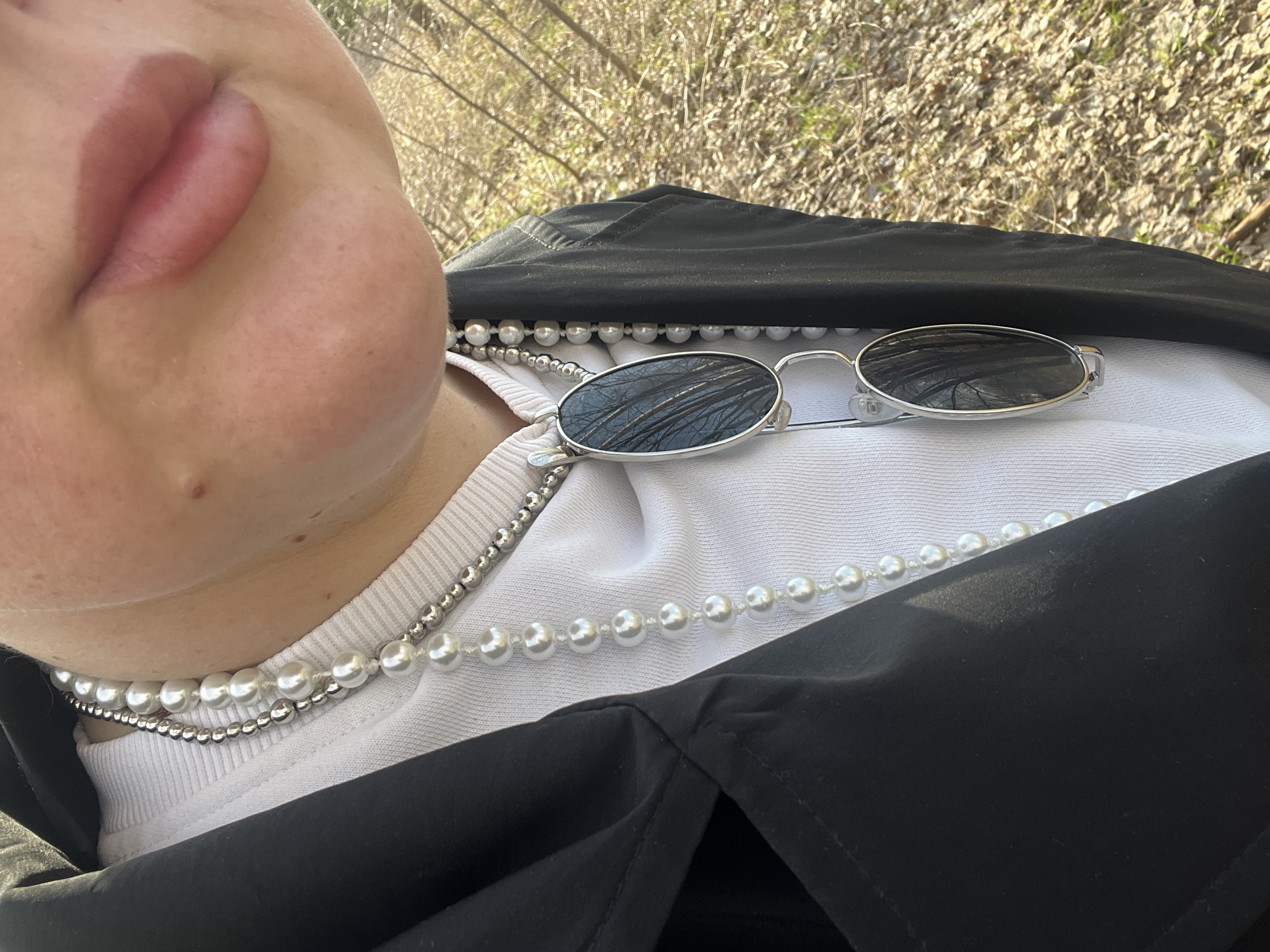 A close-up, angled shot focuses on a person's face, specifically the lips and lower left portion of the face, alongside the neck and chest area. The person is wearing two necklaces: one consisting of large, white pearls and another of smaller, silver-colored beads. They are also wearing a white t-shirt underneath a black jacket. On top of the jacket and shirt, a pair of silver-framed sunglasses rests. The background appears to be an outdoor area, possibly a park, suggested by the blurry presence of trees and ground cover. The photo's location is identified as Chaikovsky, Russia.