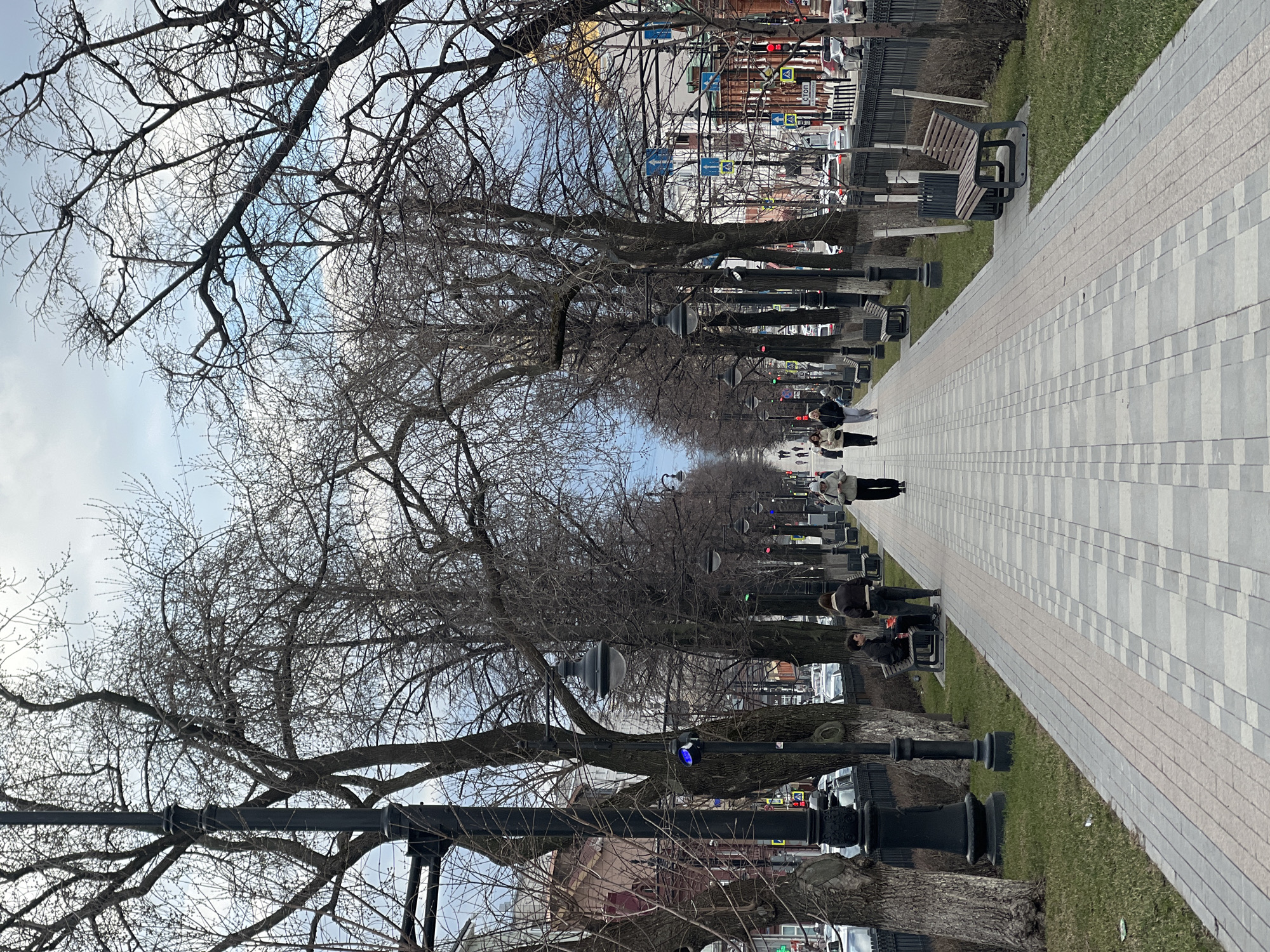 The image shows a pedestrian walkway lined with bare trees in Perm, Russia. The walkway, composed of light gray and beige pavers, is bordered by grass on one side and a line of trees and lampposts on the other. Several benches are situated along the walkway. People are visible walking and sitting on the benches. Beyond the trees, a street with vehicular traffic and buildings is visible. The sky is overcast.