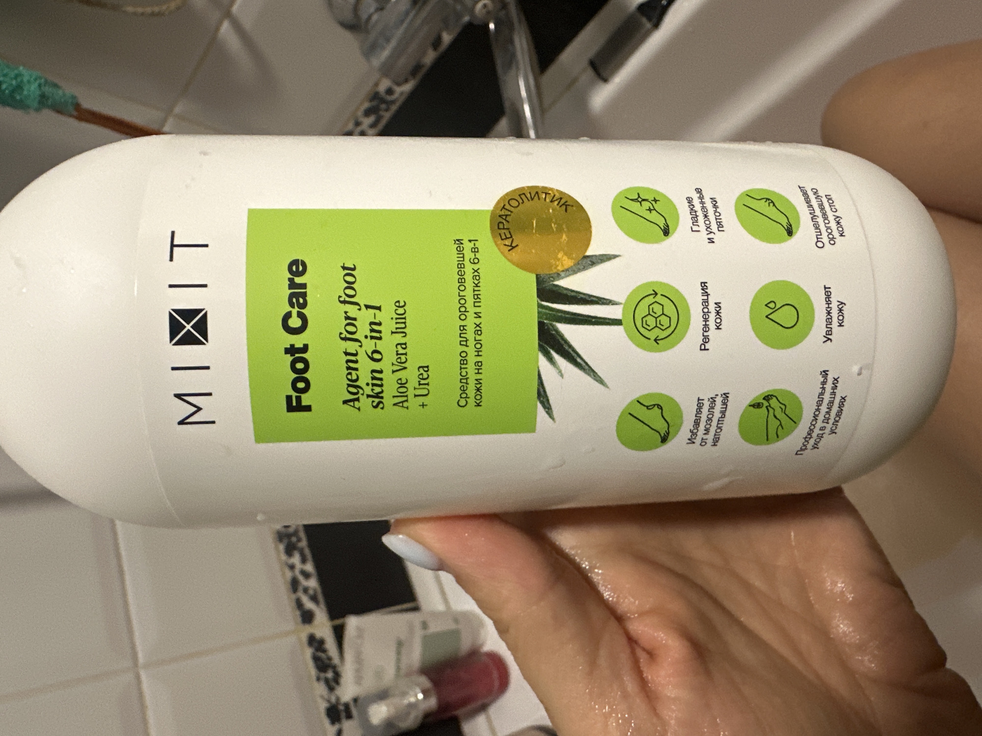 An individual is holding a white bottle of "MI&IT Foot Care Agent for foot skin 6-in-1 Aloe Vera Juice + Urea" in a bathroom. The bottle has a green label with text in English and Russian, and several circular icons depicting benefits of the product. The background shows a tiled wall and what appears to be a bathtub. The location of Novoaltaysk, Russia is not depicted in the image.