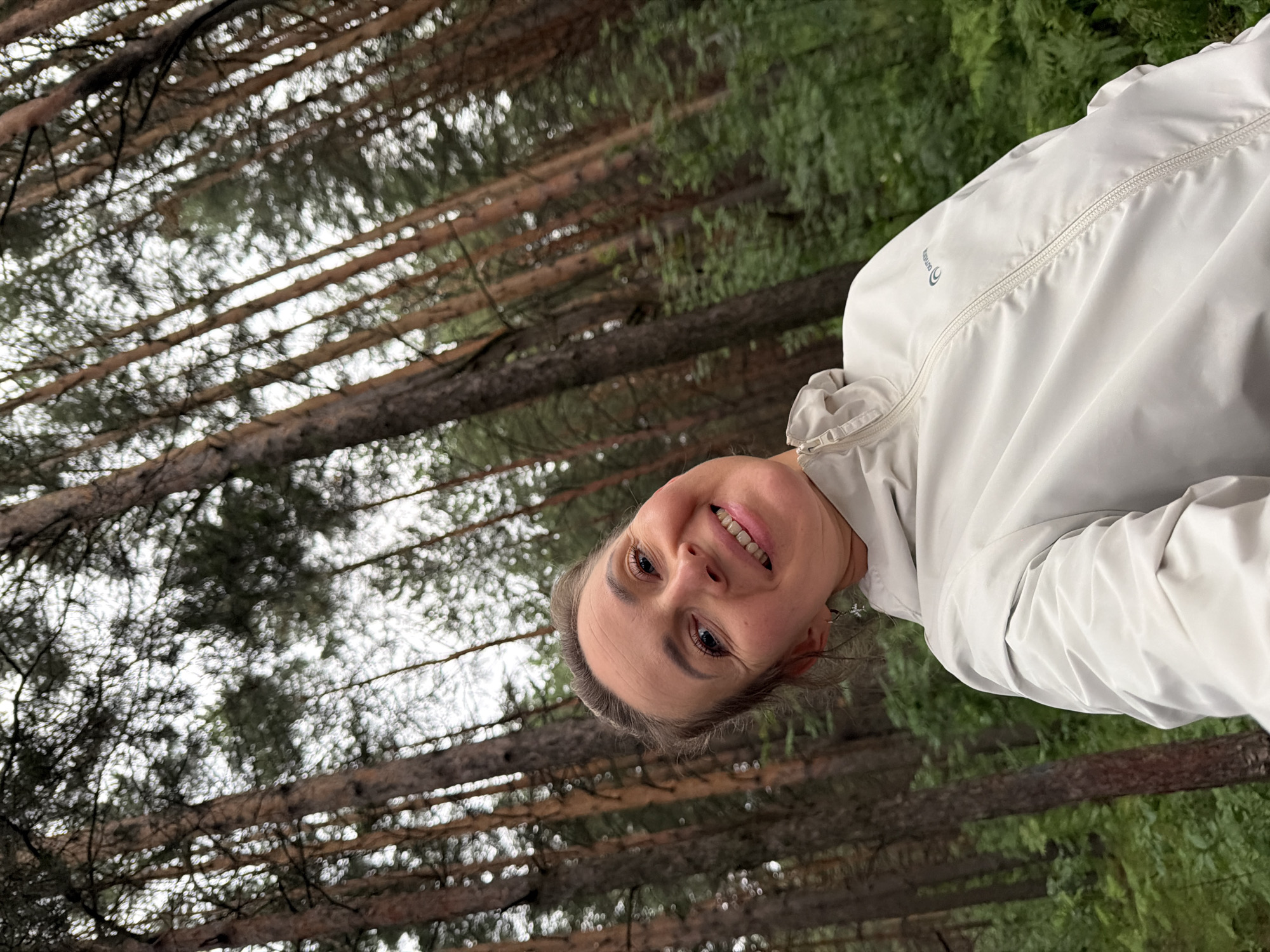 This is a selfie taken outdoors in a forest, likely in Chaikovsky, Russia. The image is oriented sideways, with the subject's head in the upper left quadrant. The subject is a woman with a smiling expression, her head tilted towards the camera. She is wearing a light-colored, possibly white or light gray, jacket with a zipper. The background is dominated by tall, slender trees, characteristic of a pine forest, with some greenery visible. The lighting suggests it is daytime, possibly overcast, as there are no strong shadows. The overall mood appears cheerful and relaxed, as indicated by the woman's smile. There is a visible logo on the jacket, which appears to be text and a circular symbol.