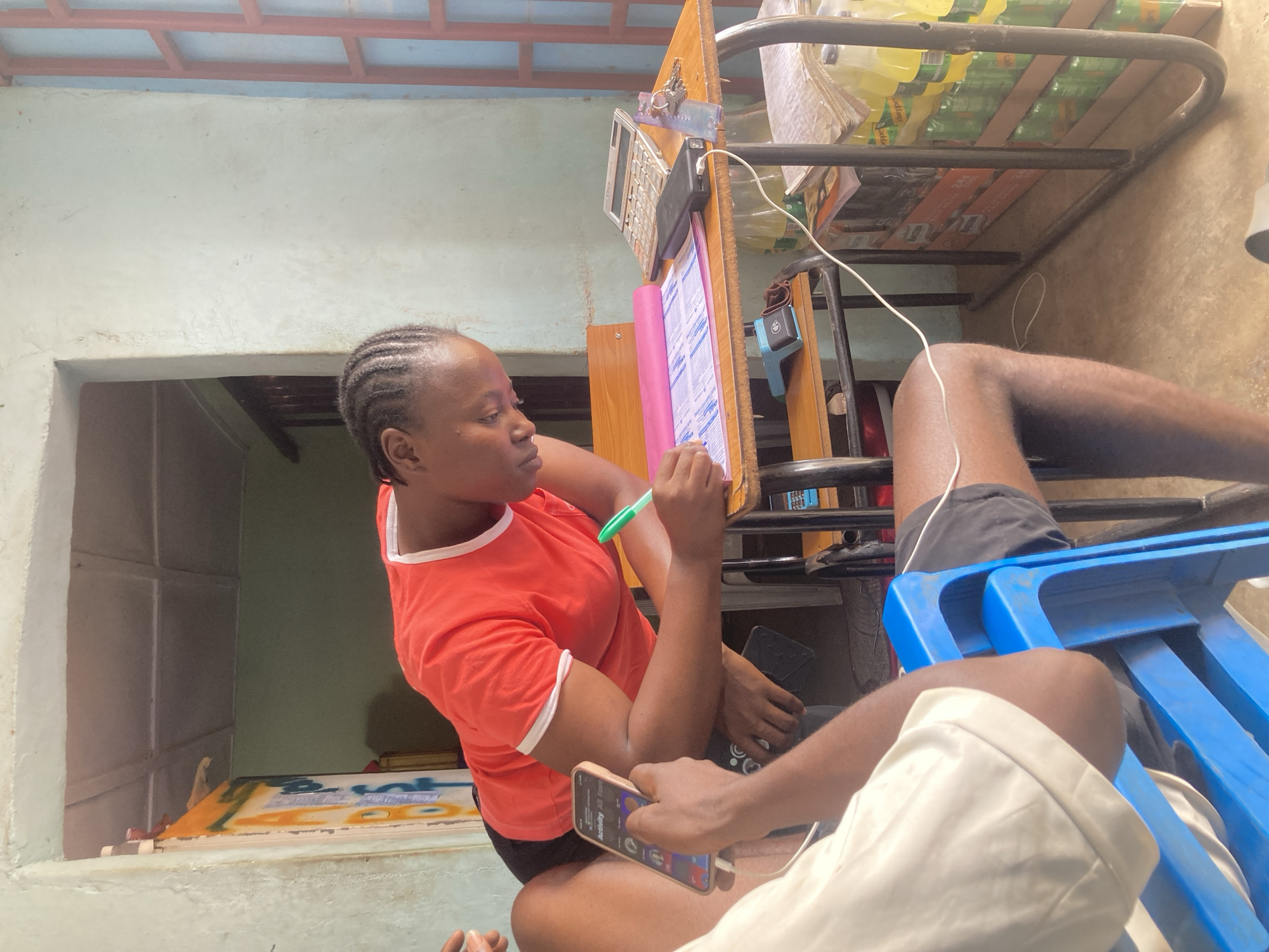 A woman sits at a desk, marking a paper with a green pen. She wears an orange shirt with white trim, and her hair is braided. A calculator and other items are on the desk, along with a carton of bottled beverages stacked on a metal shelf above. In the foreground, another person is partially visible, seated in a blue plastic chair and holding a smartphone. The scene appears to be indoors, with a doorway visible in the background. There is no evidence to confirm that this is in Jos, Nigeria.