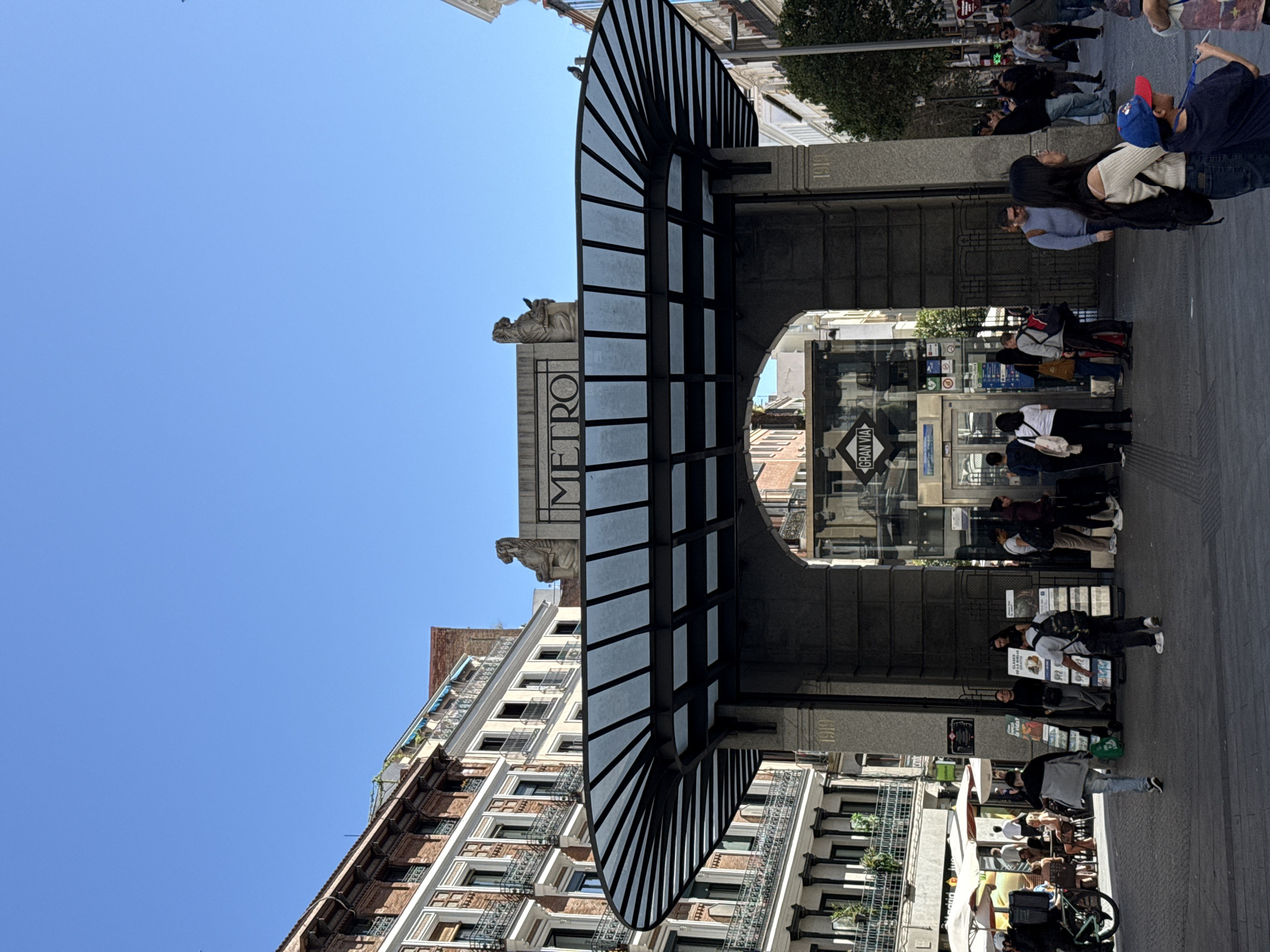 This image shows the Metro entrance at Gran Via in Madrid, Spain. The Metro entrance features a stone structure with the word "METRO" inscribed vertically. The Metro symbol is on top and two sculptures of griffins are on either side. The station’s sign that says “Gran Via” can be seen through the entrance. A canopy with metal ribbing and glass panels extends over the entrance. The building next to the structure is tall, with multiple floors, windows, and balconies. People are walking on the sidewalk near the Metro entrance, and some appear to be waiting or looking at something. There are outdoor tables set up along the street with people sitting at them. The sky is clear and blue.