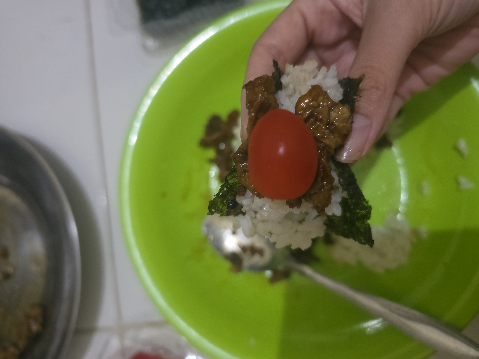 A hand is holding a small rice ball or onigiri topped with a red cherry tomato. The rice ball also contains nori seaweed and some cooked meat. The onigiri is being held over a bright green bowl, which contains more rice and bits of meat and seaweed. A spoon is also in the bowl. In the background, there's a metal bowl containing more food. The image appears to be taken in a domestic kitchen environment. The location, Pintomakmur, Indonesia, is stated in the instructions but not evident from the image content.
