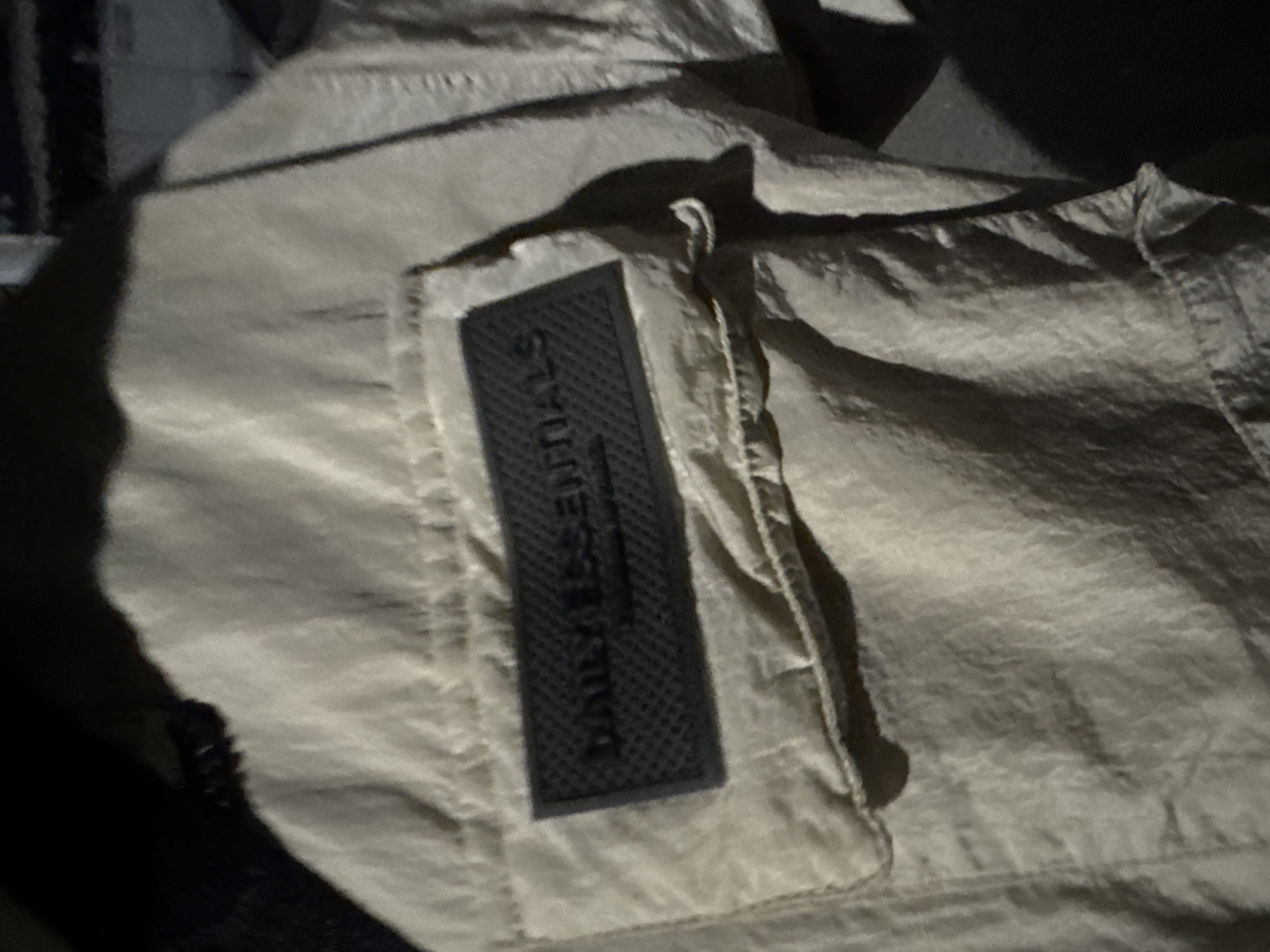 The image shows a close-up of a beige fabric garment, possibly a jacket, with a dark rectangular patch bearing the word "ESSENTIALS" in capital letters. The fabric has a crinkled texture. The background is dark and indistinct. The location is not discernible from the image.