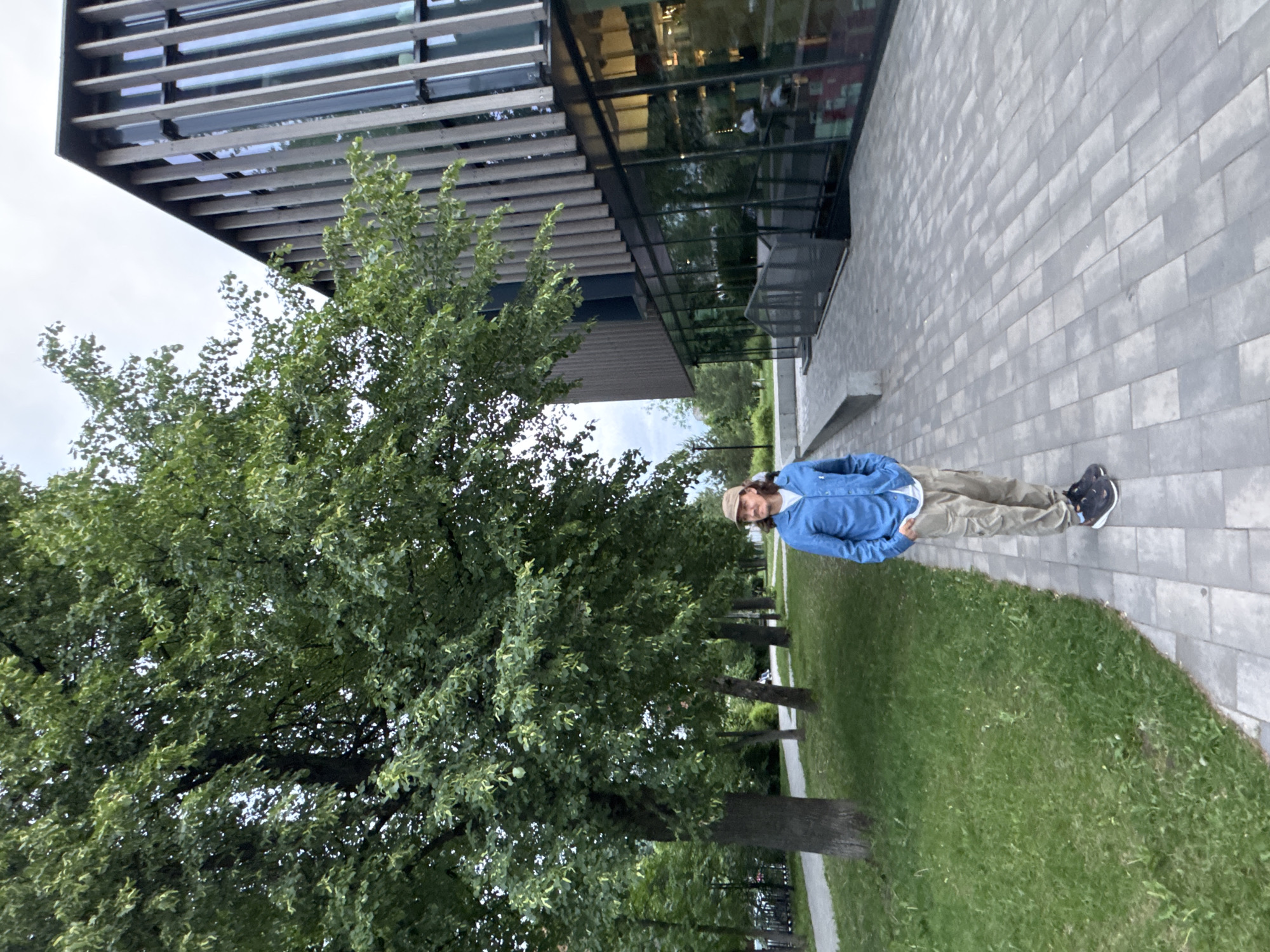 A male individual stands on a grey paved walkway, facing the right side of the frame with his head turned towards the left. He has long brown hair and wears a beige baseball cap, a light blue long-sleeved button-up shirt over a white t-shirt, khaki cargo pants, and dark athletic shoes with white soles. His hands are in his pockets, and he displays a neutral expression. To his left, a lush green grassy area is bordered by a row of mature trees with dense green foliage, extending into the background where another narrower paved path is visible. To his right, a modern multi-story building with a distinctive architectural design features a dark lower section with extensive glass windows reflecting the environment. The upper stories are characterized by numerous vertical, light-colored wooden or metallic slats that create a textured facade. The sky is overcast, indicating soft, diffused lighting. The scene is located on the campus of the Izhevsk State Technical University (ISTU) named after M.T. Kalashnikov in Izhevsk, Russia, identifiable by the distinctive modern architecture of one of its academic or research buildings.