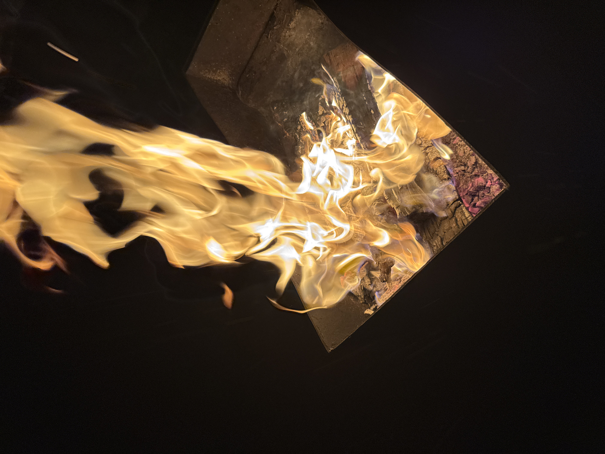 The image shows a fire burning inside a dark, rectangular container. Flames are exiting the container, extending towards the left side of the image. The container appears to hold wood and other fuel. The location of Mayak, Russia, is not directly discernible from the image. The scene depicts a fire within an enclosed space, with flames extending outward.