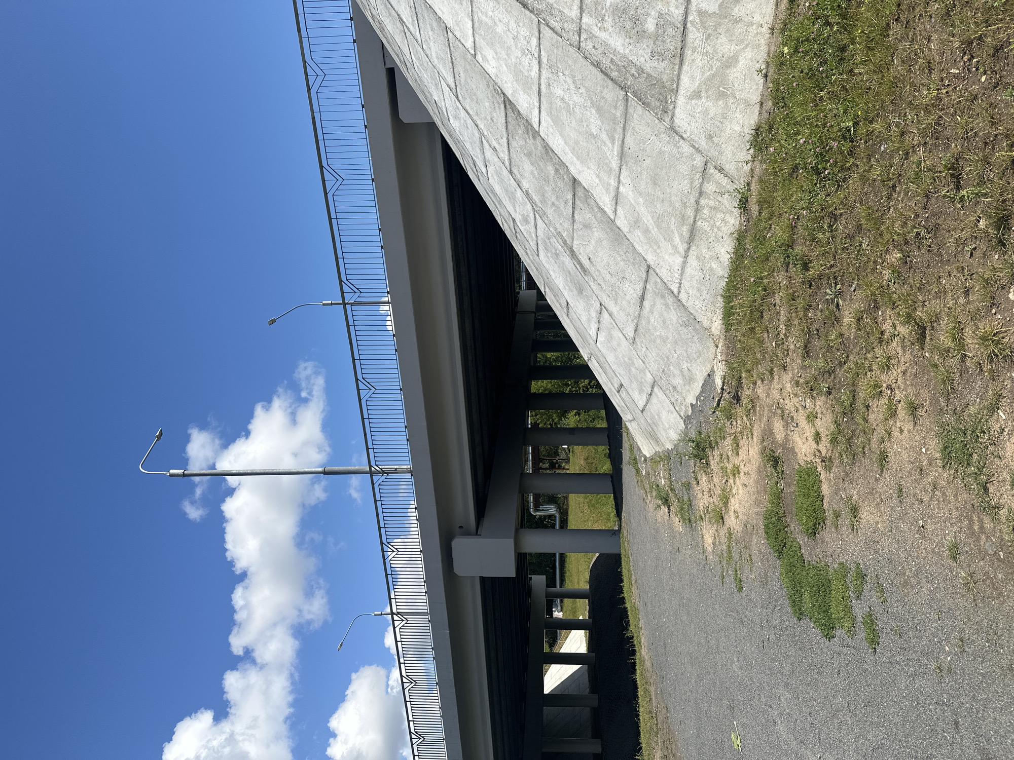 A concrete overpass stretches across a bright blue sky dotted with fluffy white clouds. The overpass has a metal railing along its top edge, and two streetlights are visible. The underside of the overpass is supported by multiple concrete pillars. In the foreground, a gravel path runs alongside a grassy area with a large, sloping concrete embankment covered in rectangular slabs. Trees are visible behind the overpass. The scene appears to be located in an urban or suburban area, possibly on the outskirts of Minsk, Belarus, given the architectural style and landscaping. There are no people visible in the image.