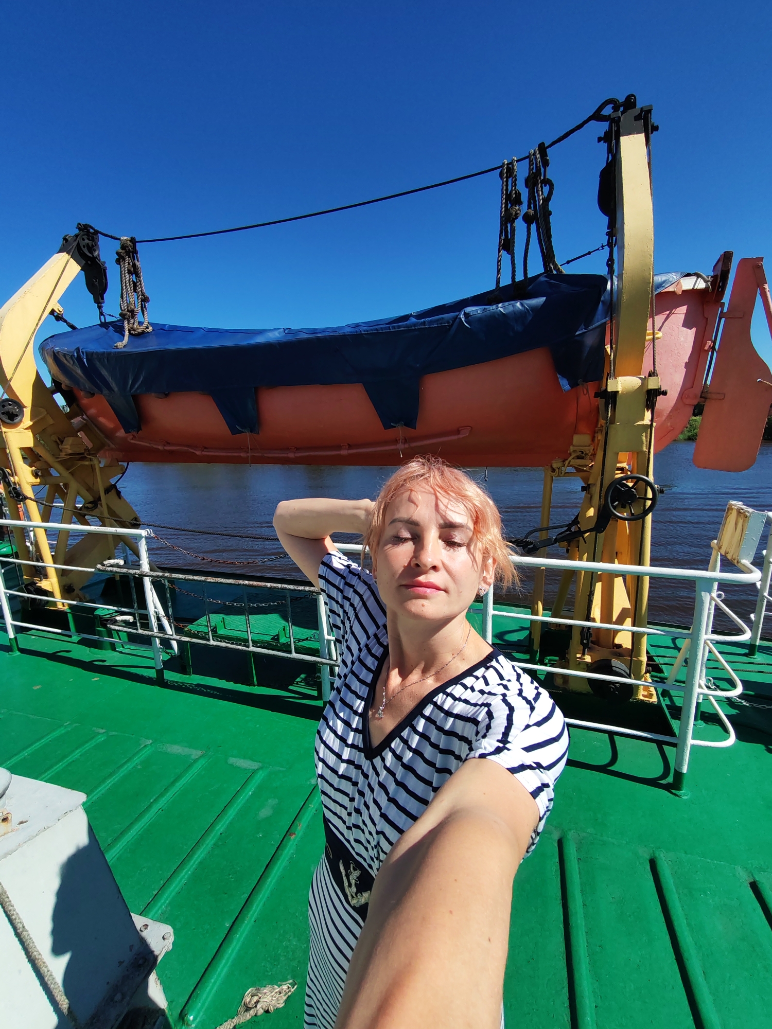 A woman with short, pink hair, wearing a black and white striped dress, is taking a selfie on the deck of a ship. She has her right arm raised, with her hand behind her head. Behind her, a lifeboat is suspended from a yellow davit against a clear blue sky. The deck is painted green, and there are white railings. The scene appears to be located in the city of Vytegra, Russia, with the Onega Canal visible in the background.