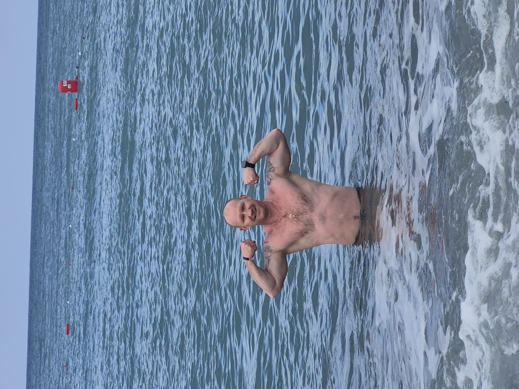 A man stands waist-deep in the sea, flexing his arms and smiling. He is shirtless, wearing a black watch on his left wrist and a necklace with a cross pendant. The water is a light blue color with small waves, and there is some white foam near the shore. In the background, there is a line of red buoys in the water. The location is Alanya, Turkey, as evidenced by the buoys marking off a swimming area in the Mediterranean Sea.
