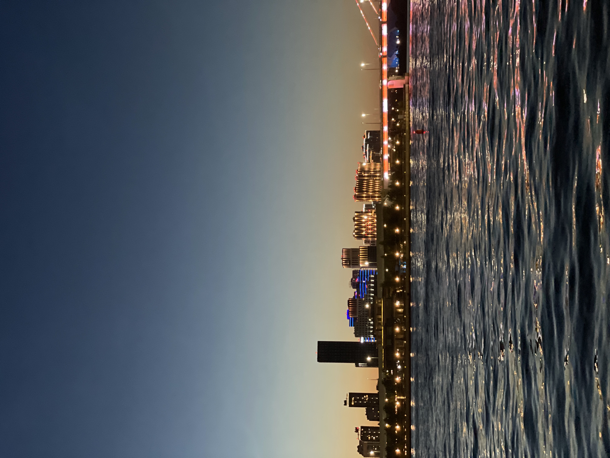This image captures a cityscape of Kazan, Russia, at dusk. A body of water, likely a river or a bay, occupies the foreground, its surface rippled and reflecting the lights of the city and the bridge. In the background, a skyline of buildings is illuminated. The tallest structure is a dark rectangular skyscraper in the center. To its left are several other buildings of varying heights, some with illuminated windows. To the right of the skyscraper, a cluster of buildings feature decorative lighting in blue and pink hues, with one building displaying a facade with a diamond pattern illuminated in orange. A bridge with orange lighting spans across the scene to the right, with cables visible. The sky transitions from a deep blue at the top to a pale orange near the horizon. Trees are visible along the waterfront in front of the buildings. There are no people or animals visible in the image.