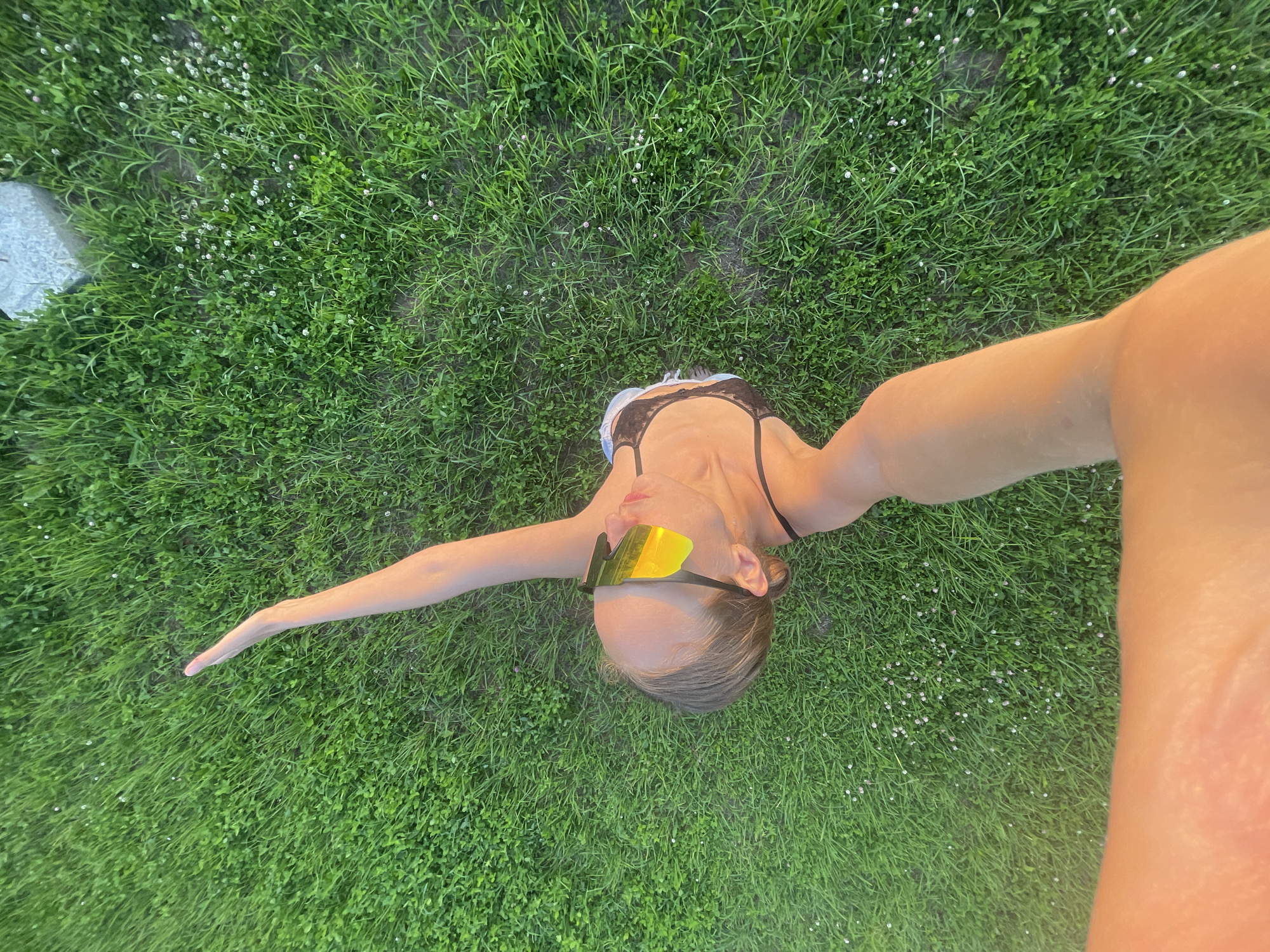 A high-angle shot shows a person lying on a patch of green grass, possibly in a park or yard in Podshivalovo, Russia. The person is wearing a black bikini top and bright yellow mirrored sunglasses. Their left arm is extended upwards and to the left. The grass is lush and green, with some small white flowers scattered throughout. A grey stone is visible in the upper right quadrant of the image. The lighting suggests it is late afternoon or early evening, with warm sunlight casting a golden hue on the person's arm.