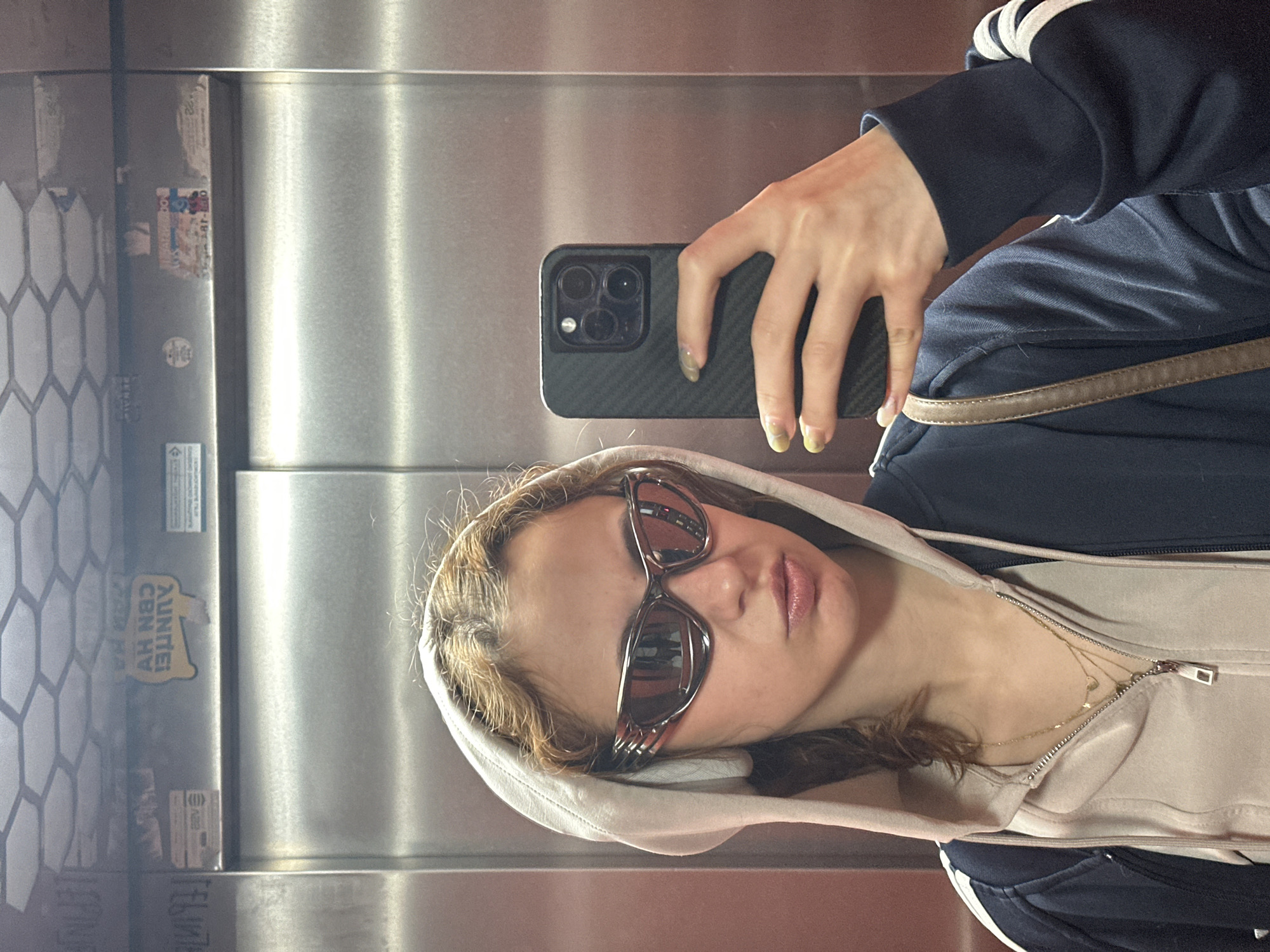 A woman is taking a selfie in an elevator. She is wearing sunglasses and a hooded sweatshirt. She is holding a phone in her right hand, with her hand slightly rotated to show off the phone’s camera lenses. She is wearing a dark blue tracksuit jacket with white stripes on the sleeve. The elevator's interior is stainless steel. A sticker with Serbian writing "AUNTEE! CBN HV" is visible on the left side of the frame. The city of Novi Sad, Serbia, is deduced from the sticker's Cyrillic script.