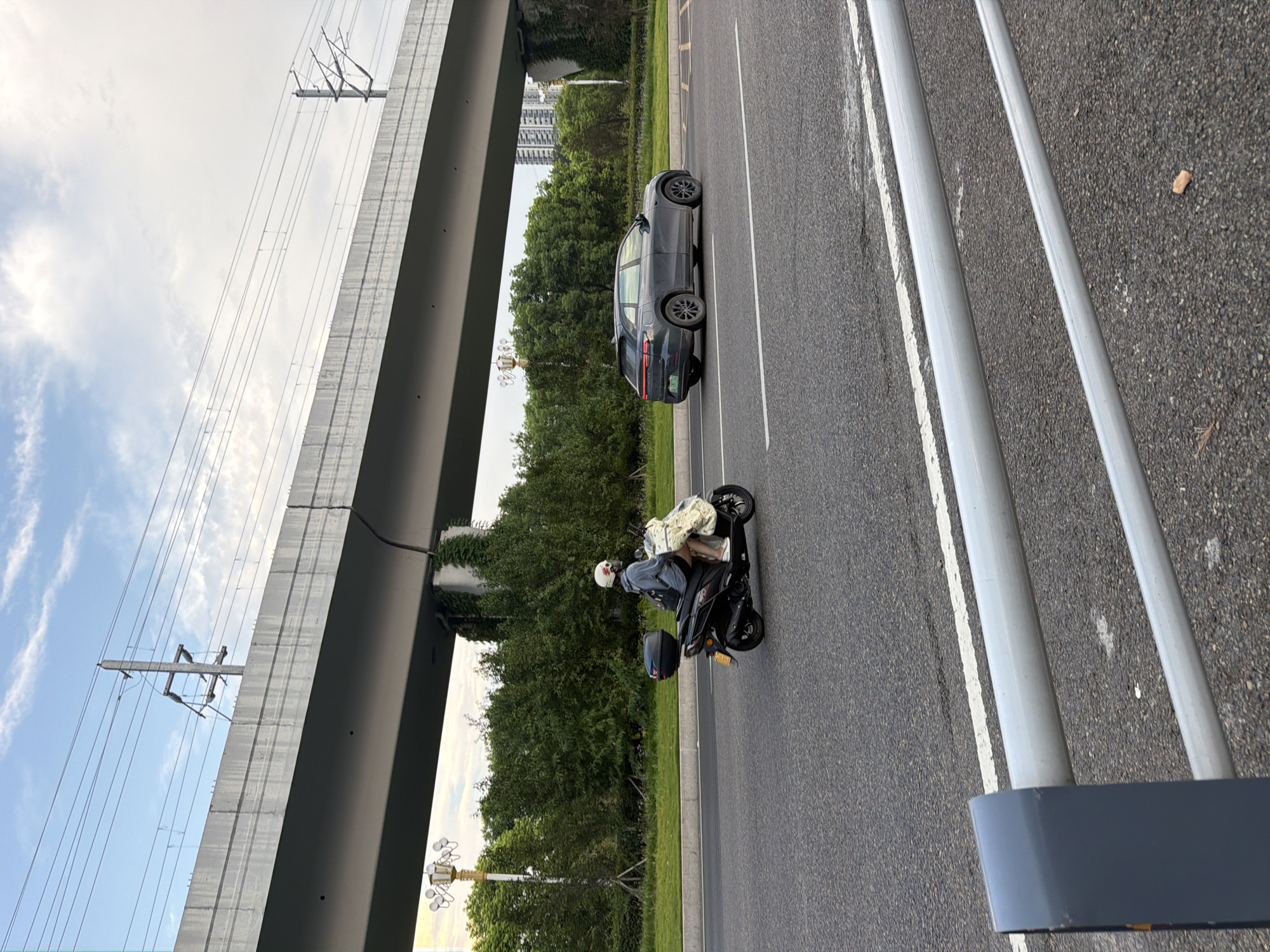 An elevated highway overpass, supported by concrete pillars entwined with greenery, casts a shadow over a multi-lane asphalt road in Jiangjin, China. Power lines crisscross the pale blue sky above the overpass. On the road, a person wearing a helmet rides a black scooter, carrying a large white bag behind them. A dark gray SUV is traveling in the adjacent lane. The foreground features a metal guardrail with gray posts. The scene captures a moment of urban transit with elements of infrastructure and nature coexisting.
