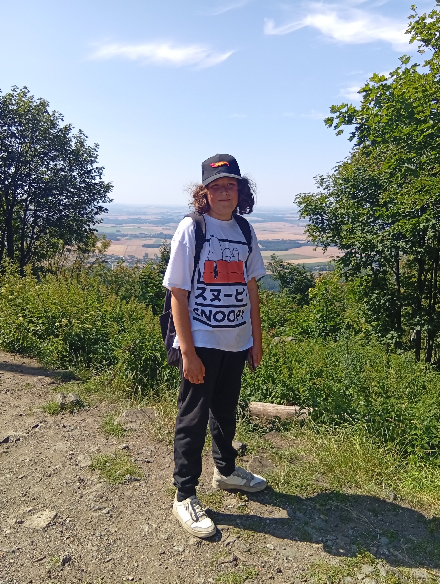 A young person stands on a dirt path, facing the camera, with a scenic view of fields and trees in the background. The person is wearing a black baseball cap, a white t-shirt with a graphic print, black pants, and white sneakers. A black backpack is visible over their shoulders. The path is rocky and dusty, with green grass and shrubs growing alongside it. In the background, rolling hills and farmland stretch towards the horizon under a clear blue sky with scattered clouds. The location is identified as being in the city of Sobótka, Poland.