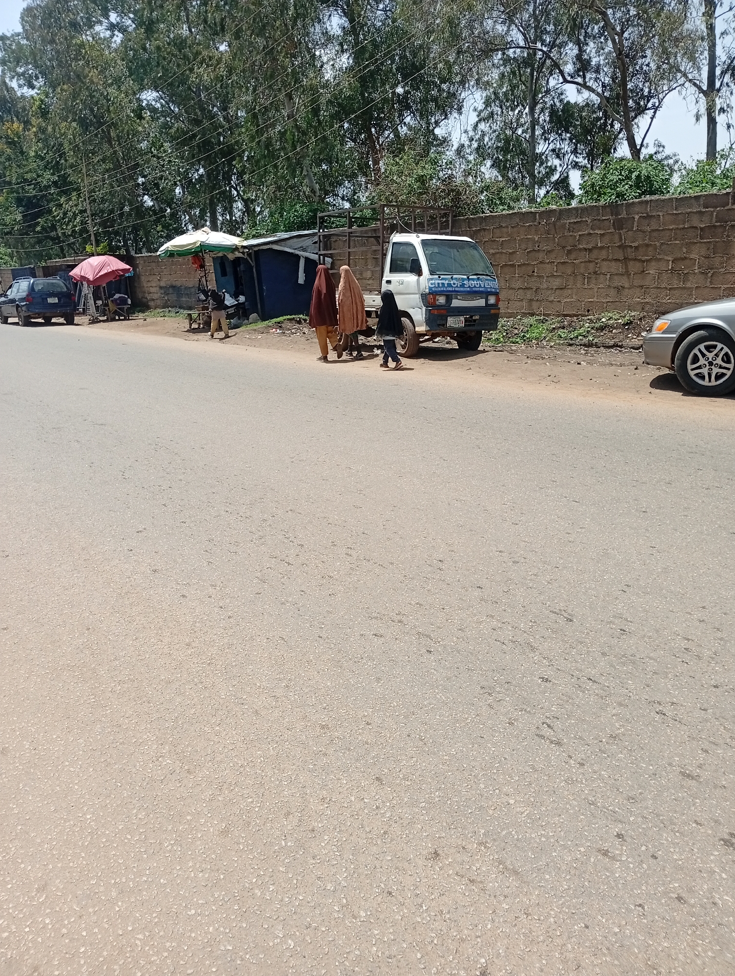 A scene unfolds along a paved road in Jos, Nigeria, marked by a brick wall running parallel to the street. In the distance, several trees stand tall, their foliage creating a natural backdrop. A small truck with "CITY OF SOUVENIRS" printed on its side is parked near the wall, its white body contrasting with the earthy tones of its surroundings. Several people are visible near a makeshift stall, some dressed in traditional attire. Two women in vibrant headscarves, one in maroon and the other in beige, are walking away from the stall. A young child in a black headscarf and dark clothing walks ahead of them, seemingly on their way to a different part of the scene. In the foreground, a silver car is parked on the side of the road, its rear tire visible, suggesting a bustling environment where people and vehicles coexist.