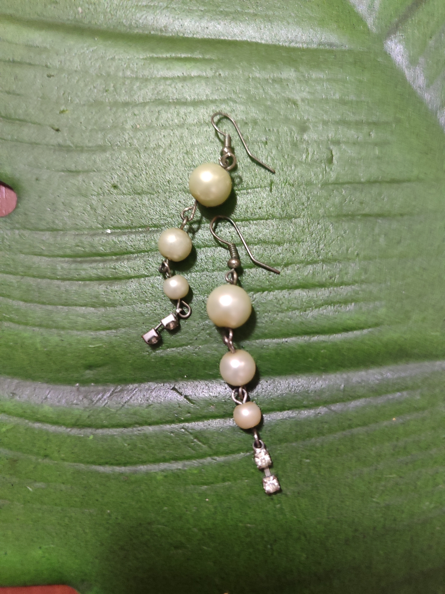 A pair of pearl and rhinestone earrings is displayed on a green leaf. The earrings are made of silver-colored metal and feature multiple pearl beads of varying sizes. One earring has a dangle with three pearls and two square rhinestones at the bottom. The other earring has a similar dangle with three pearls and a rectangular rhinestone embellishment. The background is a textured green leaf with visible veins. The image is taken from a top-down perspective, and there is no indication of any specific location within the city of Mykolaiv, Ukraine.