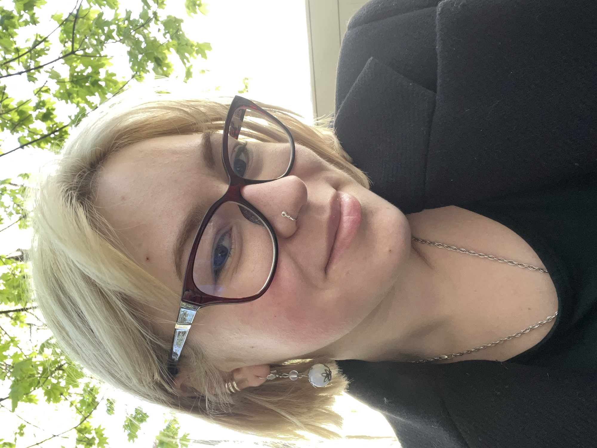 The media file features a close-up selfie of a young woman taken outdoors, likely in Odesa, Ukraine. The image is oriented sideways, with the woman looking directly at the camera. She has blonde hair with bangs, fair skin, and blue eyes, partially obscured by prominent dark red, cat-eye style eyeglasses. Her expression is neutral with a slight hint of a smile.
Notable facial features include a small silver nose ring and naturally pink, slightly parted lips. Her left ear (viewer's right) is adorned with two simple silver hoop earrings and a longer dangling earring featuring a large, spherical white or clear bead with a dark pattern, accompanied by smaller clear beads. A simple silver chain necklace is visible around her neck and collarbone.
She is wearing a dark-colored, possibly black, textured jacket or coat over a dark top. The background, particularly in the upper left, shows bright sunlight filtering through green leaves and branches, indicating a sunny daytime setting, possibly in a park or garden. There is no visible text in the image.