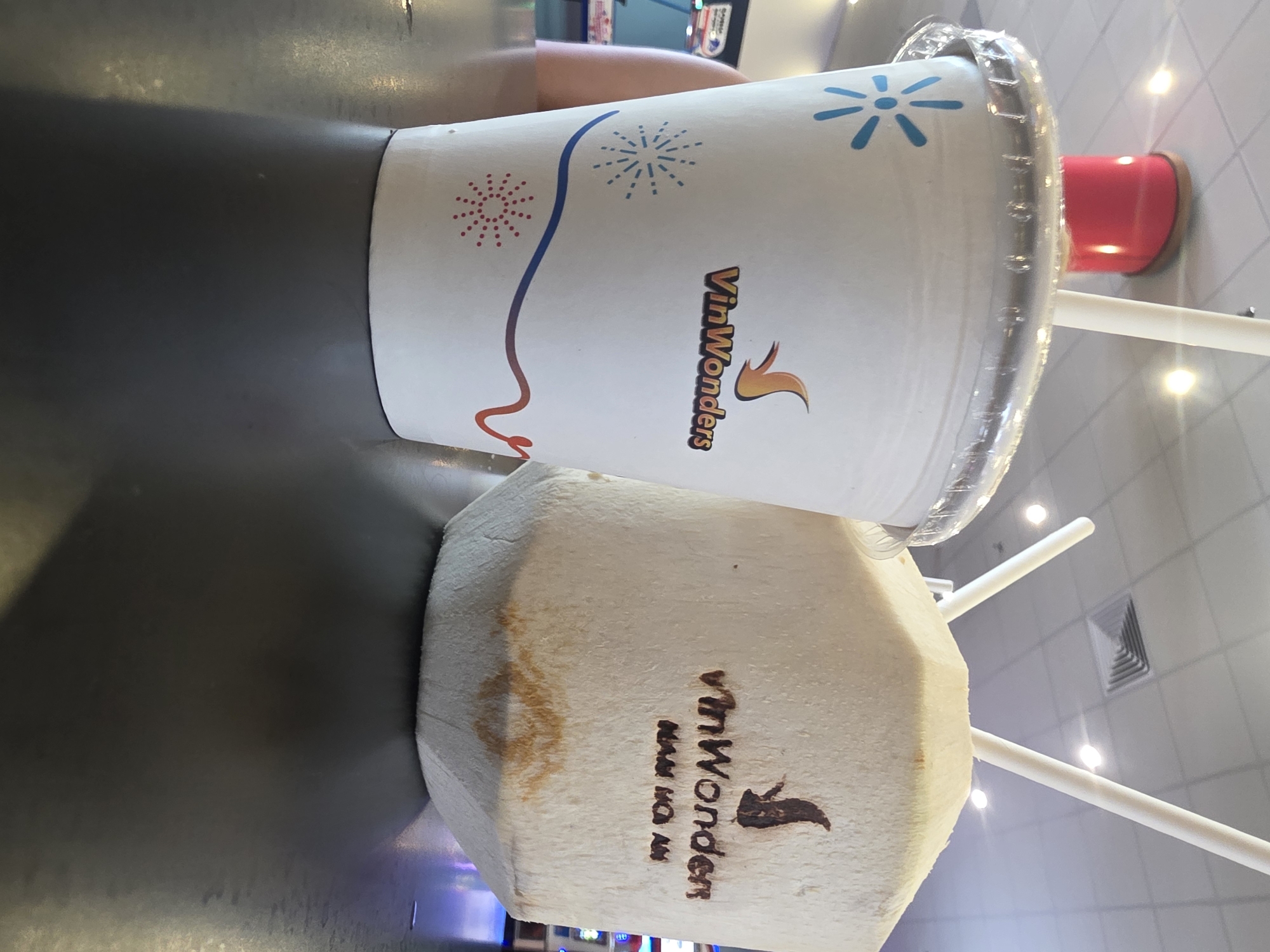 The image captures a close-up shot of a beverage cup and a whole coconut, both branded with the "VinWonders" logo, suggesting a location within a VinWonders theme park or related establishment. The beverage cup is white with a clear plastic lid, decorated with blue and red firework-like designs and the "VinWonders" name. The coconut is a light tan color, also bearing the "VinWonders" logo. Two white straws are visible, protruding from the coconut. The objects rest on a dark, glossy surface. Given the brand association, the location is likely within the city of Đà Nẵng, Vietnam, where VinWonders has a significant presence. The overall environment appears to be indoors, potentially a food court or refreshment area.