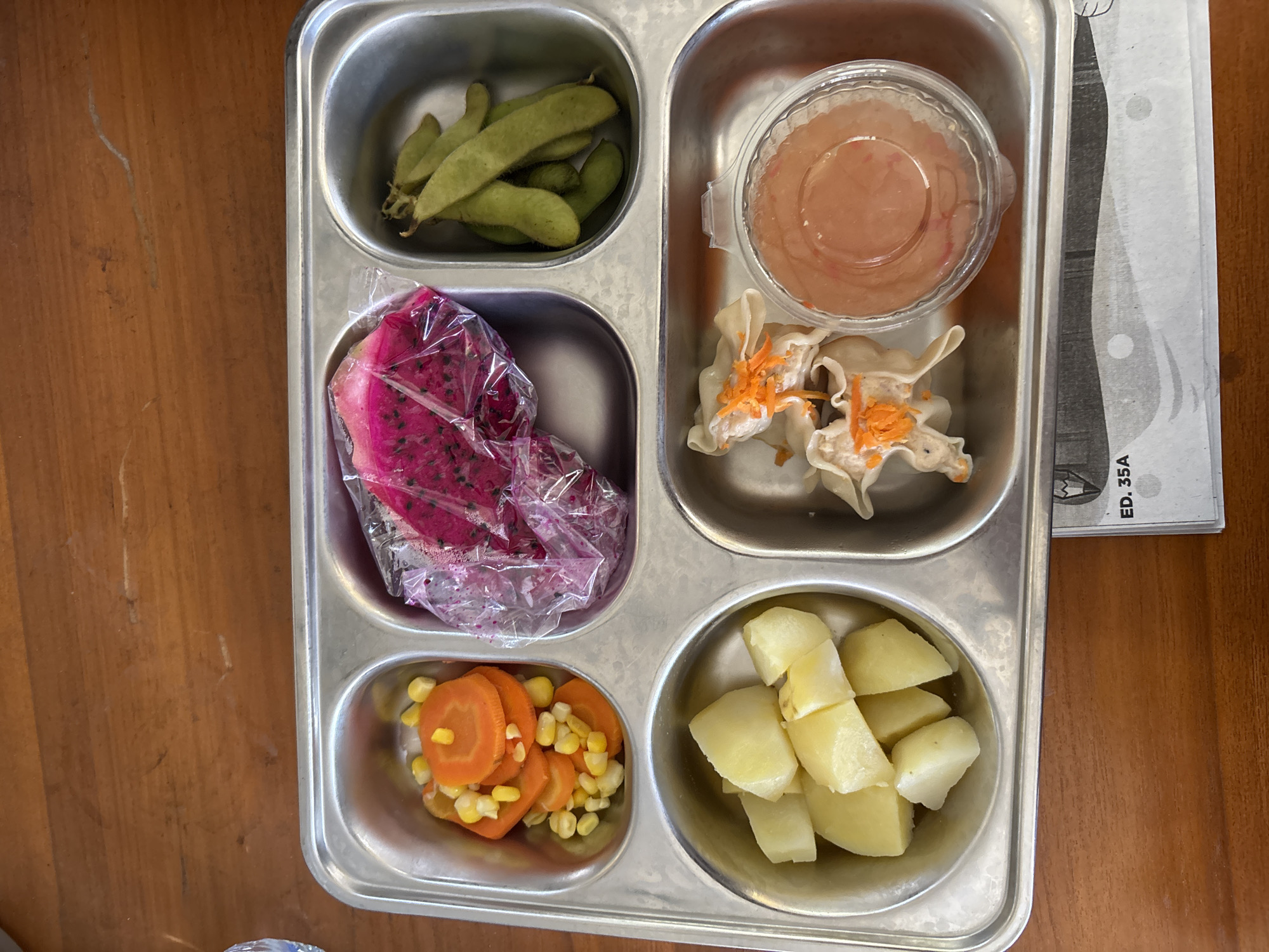 The image shows a silver compartmentalized tray filled with various food items arranged on a wooden surface. The tray contains a portion of steamed carrots and corn, sliced dragon fruit in plastic wrap, edamame pods, boiled potatoes, and two pieces of dim sum topped with shredded carrots, accompanied by a small plastic container of dipping sauce. The tray is positioned on a wooden table. In the lower right corner of the image, a piece of paper with the text "ED. 35A" is visible. The scene appears to be an overhead view of a meal. The location in Tirtomoyo, Indonesia is not evident from the image itself.