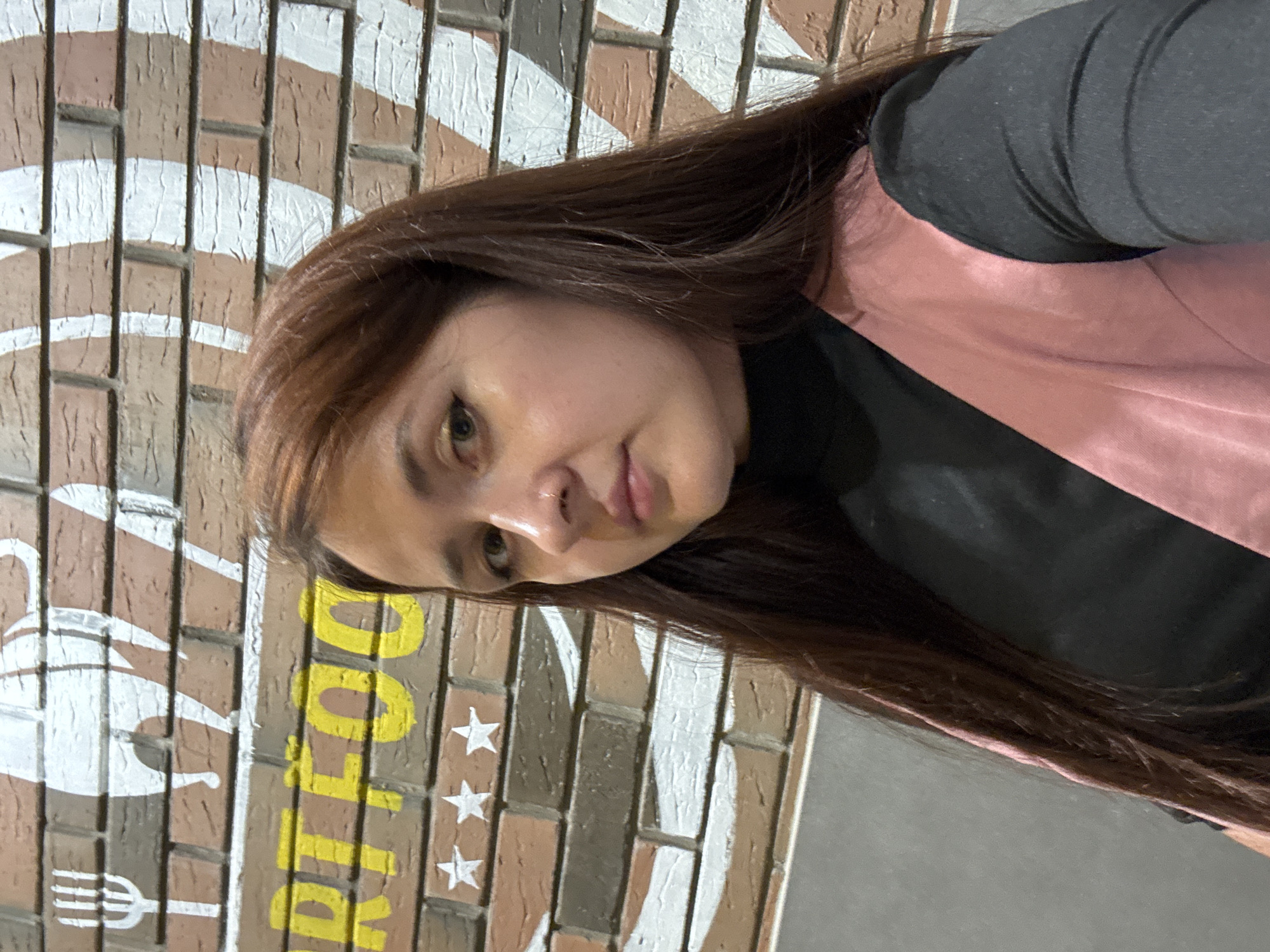 A medium shot shows a young woman with long, dark auburn hair and fair skin. She is wearing a black turtleneck and a pink vest. Her gaze is directed slightly off-camera to her right. The background consists of a brick wall with painted white and light brown stripes. Yellow lettering, partially obscured, reads "RT FOO". Three white stars are also painted on the brick wall. The lighting appears to be artificial and overhead. The image seems to have been taken indoors, possibly in a restaurant or cafe.