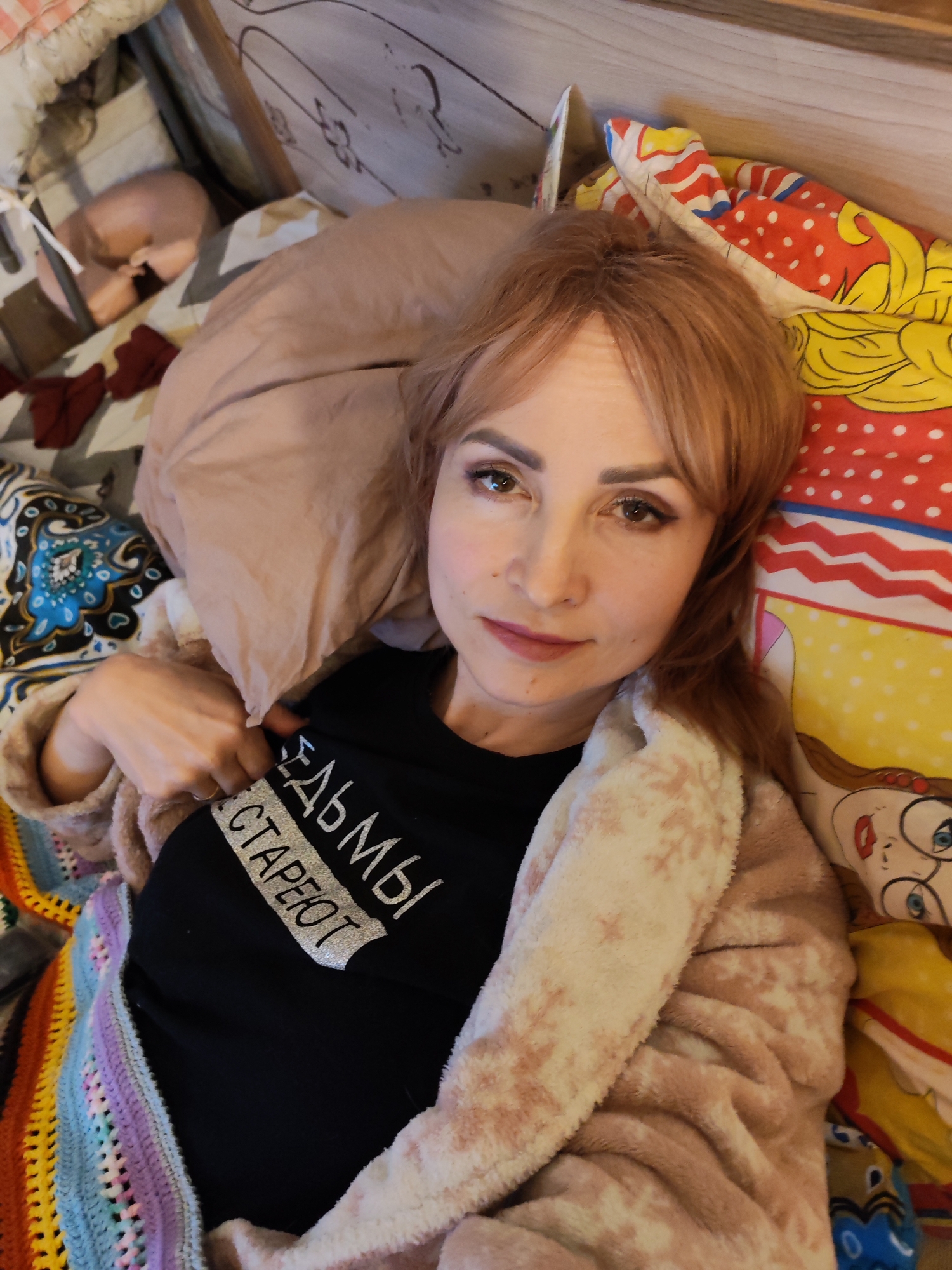 A woman with reddish-brown hair and bangs is lying on a bed, looking directly at the camera. She has light makeup, including defined eyebrows, eyeliner, and pinkish lipstick. She is wearing a black t-shirt with white Cyrillic text and a soft, pinkish-beige plush robe with white snowflake-like patterns. Her left hand is subtly pulling at the collar of her t-shirt, revealing the text.
The bed is adorned with diverse bedding. To her right, a colorful sheet with cartoon-style characters, including one resembling Minnie Mouse, is visible. To her left, there's a patterned sheet with blue, black, and white intricate designs. A vibrant, striped crocheted blanket in colors like yellow, orange, red, pink, purple, blue, and green is draped over her lower body and to her left. A plain, light brown pillow supports her head. In the upper left background, partially obscured, a small, light-colored object or animal, possibly a sphynx cat or a plush toy, is curled up. A wooden headboard with carved details is also visible behind her.
The visible text on her black t-shirt reads "ЕДЬМЫ СТАРЕЮТ". The full word is likely "ВЕДЬМЫ", so the phrase translates to "Witches get old" or "Witches are aging". The woman's expression is calm and neutral. The scene is set indoors, likely in a bedroom in Chaikovsky, Russia.