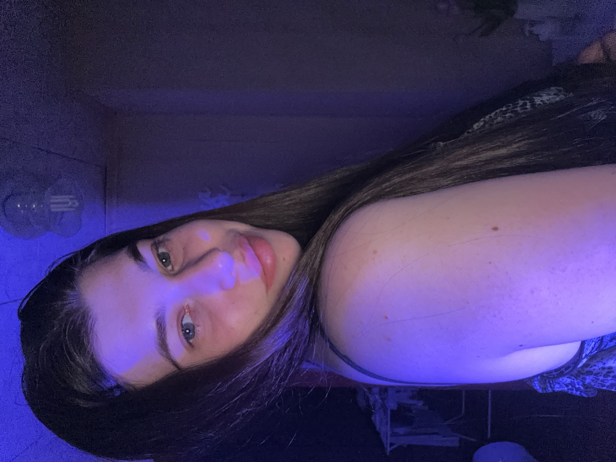 The image is a selfie of a young woman with long, dark hair. She has fair skin, light-colored eyes, and is smiling slightly. The lighting appears to be blue, possibly from an LED light source. The background is dark and indistinct, but there are hints of room furnishings, including a wall and possibly a bed or furniture. There is no information in the image indicating it was taken in Мещёры, Russia.