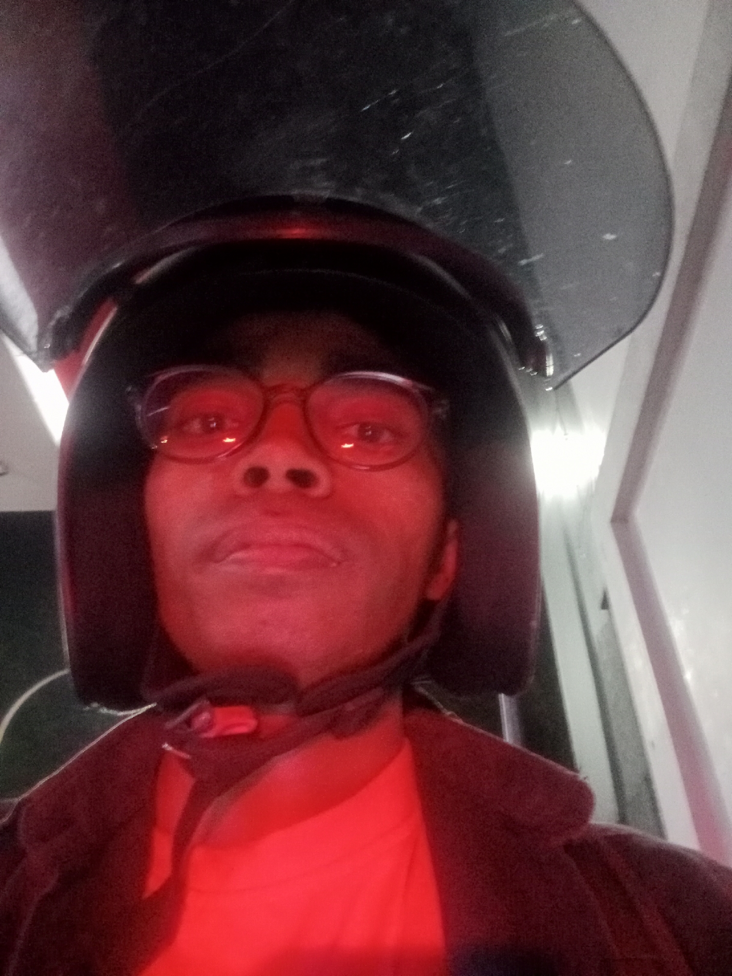 A person wearing glasses and a black motorcycle helmet with a dark tinted visor is looking at the camera. The individual is illuminated by red lighting, which casts a strong red hue across their face and clothing. They appear to be indoors, possibly in a room with white walls and a doorway visible in the background. The image does not provide any specific location details within the city of Larangan, Indonesia.