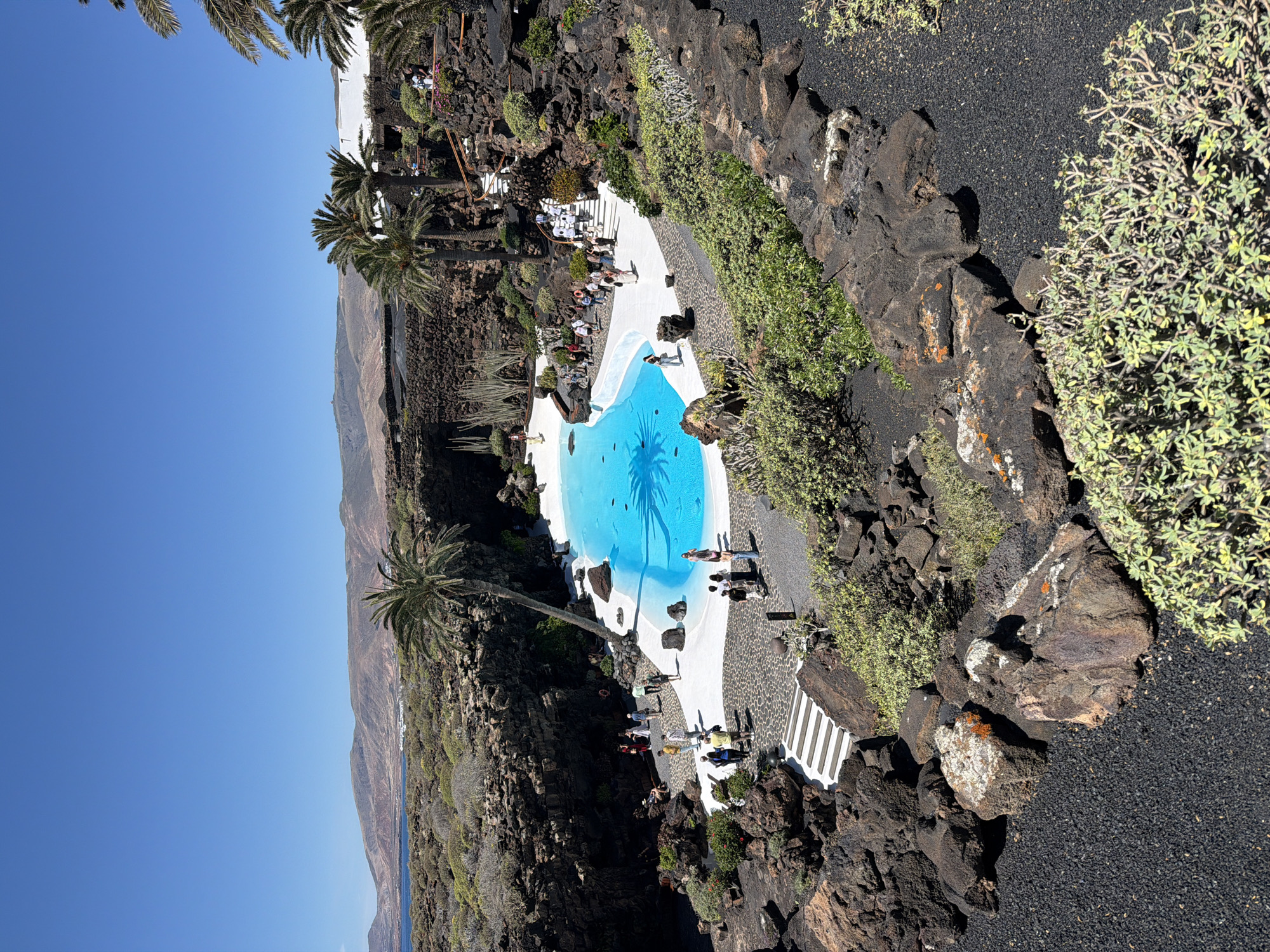The image depicts an aerial view of the Jameos del Agua in Haría, Lanzarote, Spain. It features a unique swimming pool built into a lava cave. The pool has bright blue water and white walls. Palm trees grow around the pool and cast shadows in the water. People are standing and walking around the pool area. The surrounding landscape is characterized by black volcanic rock, sparse vegetation, and a coastline with mountains visible in the distance. The sky is clear and blue.