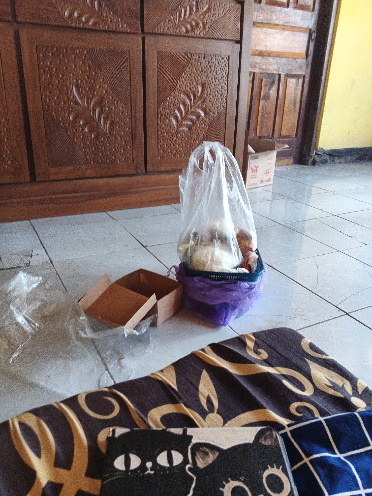In Karangjengkol, Indonesia, a bag of groceries sits on a tiled floor next to an open cardboard box. The bag, made of clear plastic, is placed inside a dark green basket lined with a purple plastic bag. Inside the plastic bag, various food items are visible, including what appears to be white rice and some reddish-brown items. To the left of the groceries, a crumpled plastic bag lies on the floor. Further to the left, an open cardboard box with its flaps extended is visible. In the background, a large, intricately carved wooden cabinet dominates the scene. To the right of the cabinet, a wooden door is partially open, revealing a section of a bright yellow wall. A small cardboard box with the "Vit" logo and text indicating a beverage is placed near the door. In the foreground, a dark brown blanket with a pattern of swirling gold shapes is spread out, partially covering a dark blue blanket with a white checkered pattern. A black object with a cartoonish cat face printed on it rests on top of the patterned blankets. The lighting suggests an indoor setting, possibly a home or a storage area.