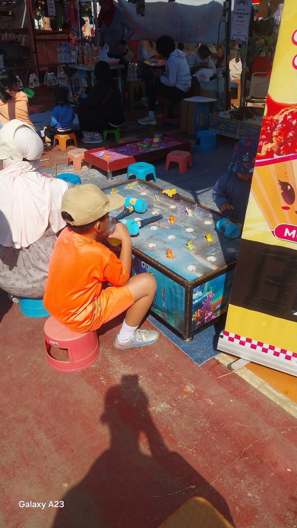 A group of children and adults are gathered around a water-themed carnival game. The game itself is a rectangular table with a blue surface designed to look like water, complete with fish and coral graphics. Several small plastic toys, including yellow ducks and what appear to be fish, are scattered across the surface. Two blue plastic hammers with white handles are resting on the table, suggesting a game where players strike targets.
In the foreground, a young boy wearing a tan baseball cap, an orange t-shirt, and orange shorts is sitting on a pink stool. He is holding a yellow cup with a straw and appears to be drinking from it. His legs are crossed, and he is looking towards the game table. He is wearing white socks and silver athletic shoes.
To the left of the boy, a person wearing a light pink hijab and a patterned dress is partially visible, also seated. To the right, a young child wearing a dark blue hat with a pattern is looking down at the game.
In the background, several other individuals are present, some standing and some seated on colorful stools. They appear to be observing or participating in activities related to the game or surrounding stalls. The scene is outdoors, with a red-colored ground surface visible. A tall, yellow and white striped banner with cartoon graphics is on the far right, partially obscuring some of the scene. The setting suggests a public gathering or event, possibly a market or festival, in Malang, Indonesia.