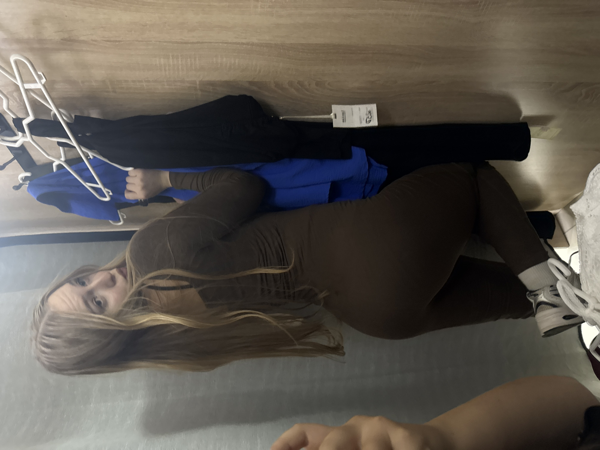 A blonde woman wearing a brown fitted jumpsuit and white socks with athletic shoes is posing in a fitting room. She is looking over her left shoulder at the camera. To her right, a rack of clothes, including a blue shirt and a black top, hangs on a white hanger. The fitting room has a wood-paneled wall on the right and a gray curtain on the left. There is a white tag hanging from the black top. The overall scene suggests a retail or personal shopping experience. There is no information in the image to indicate the location is the city of Perm, Russia.